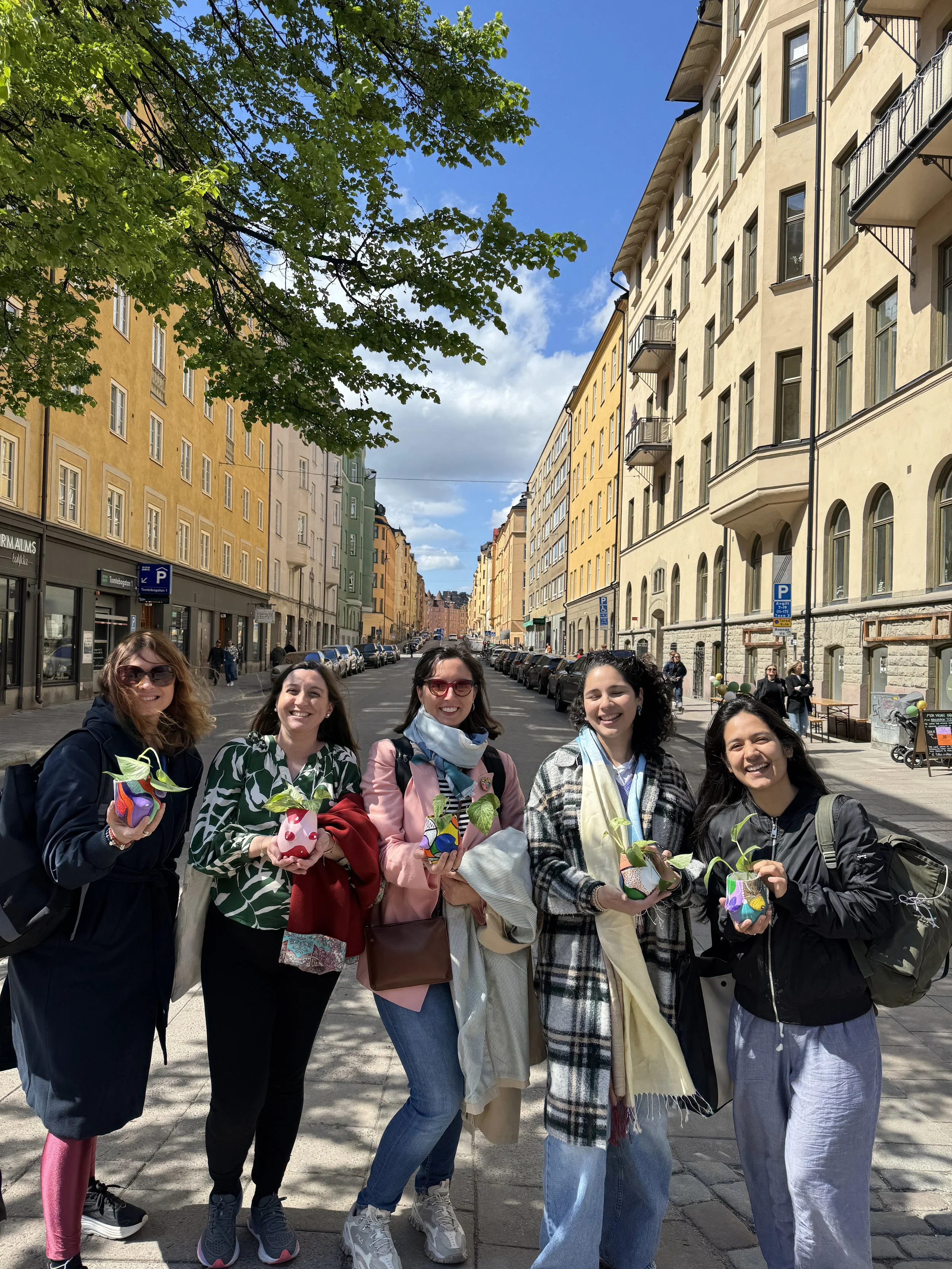 Five women standing on a city sidewalk, each holding colorful small potted plants, smiling at the camera, with yellow and beige buildings lining the street and a bright blue sky overhead.