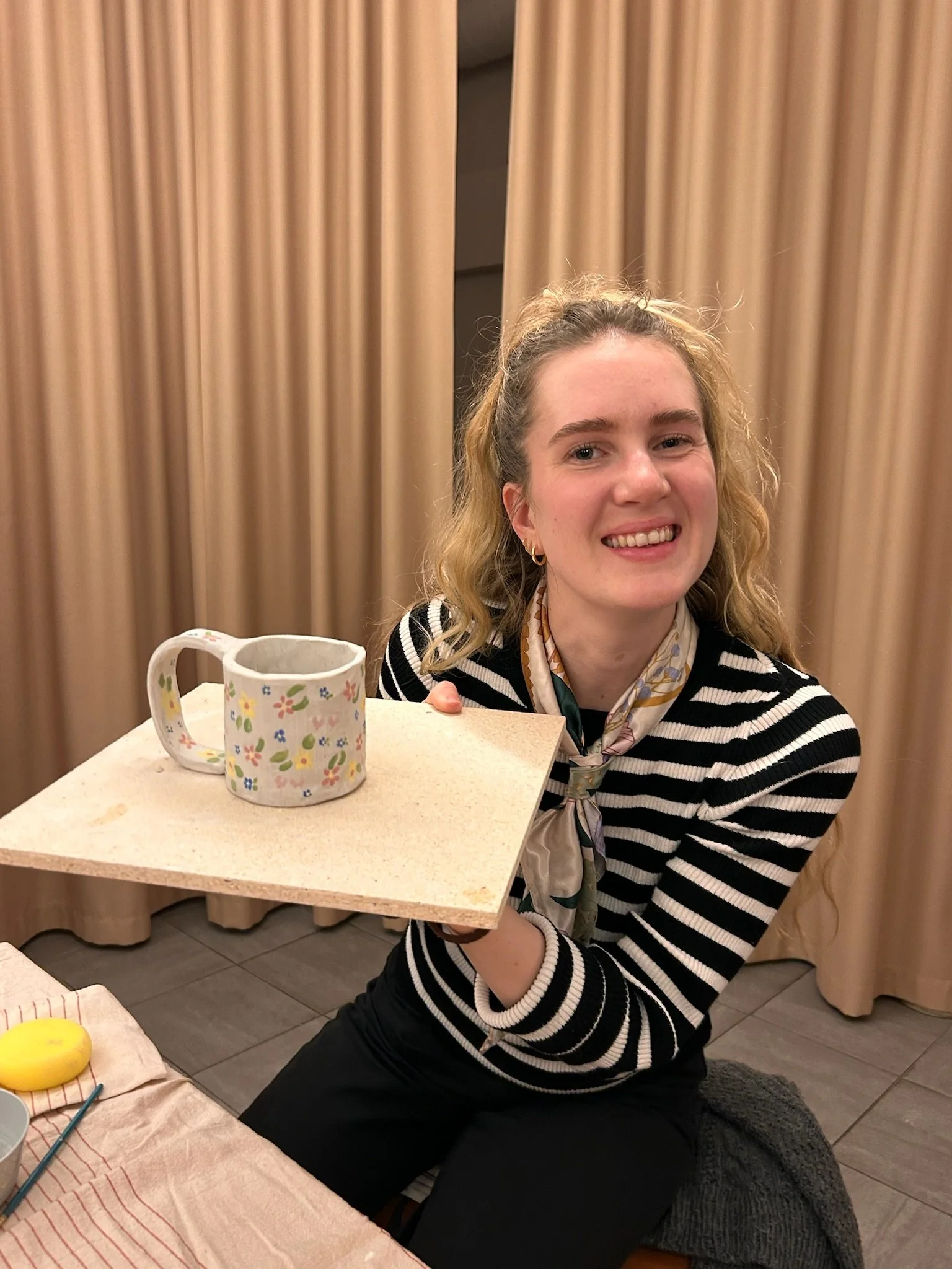 A woman with curly blonde hair, wearing a striped black and white shirt and a scarf, holding a plate with a ceramic mug on it, smiling at the camera, with beige curtains in the background.