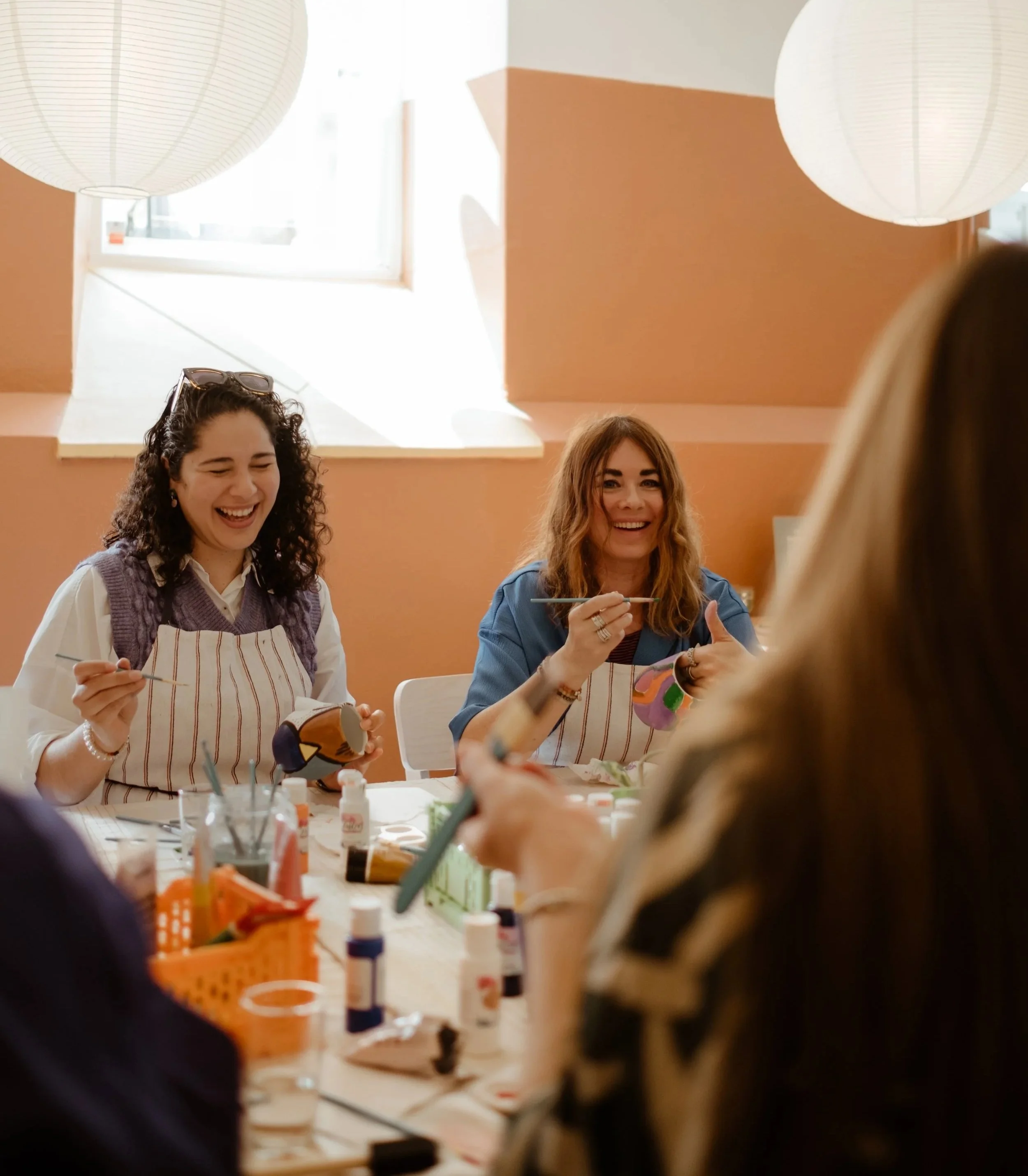 Three women sitting at a table, smiling and laughing during a crafting activity with paint supplies in a brightly lit room.