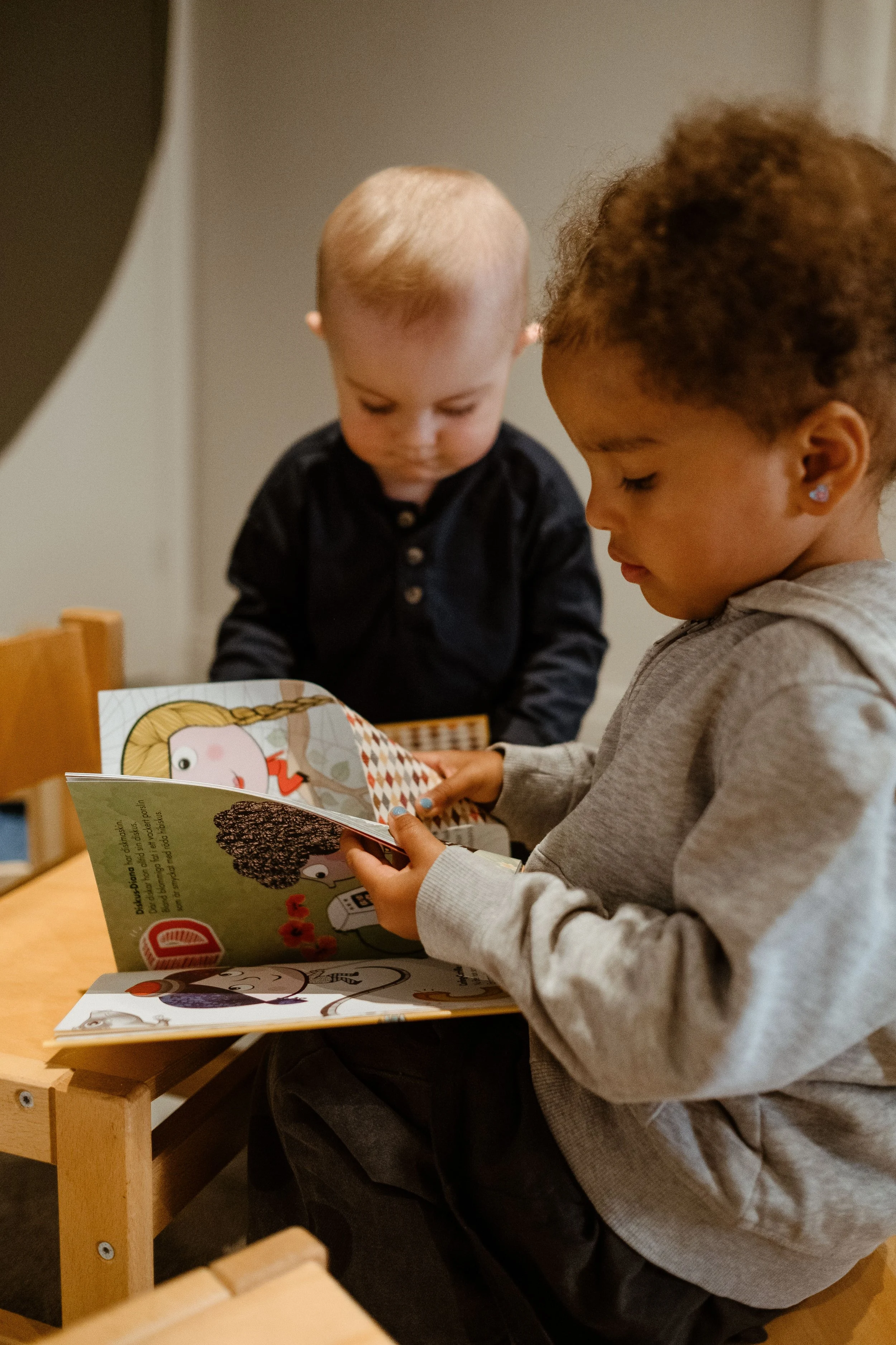 Two young children, a girl and a boy, are looking at a colorful children's book together while sitting at a small wooden table.