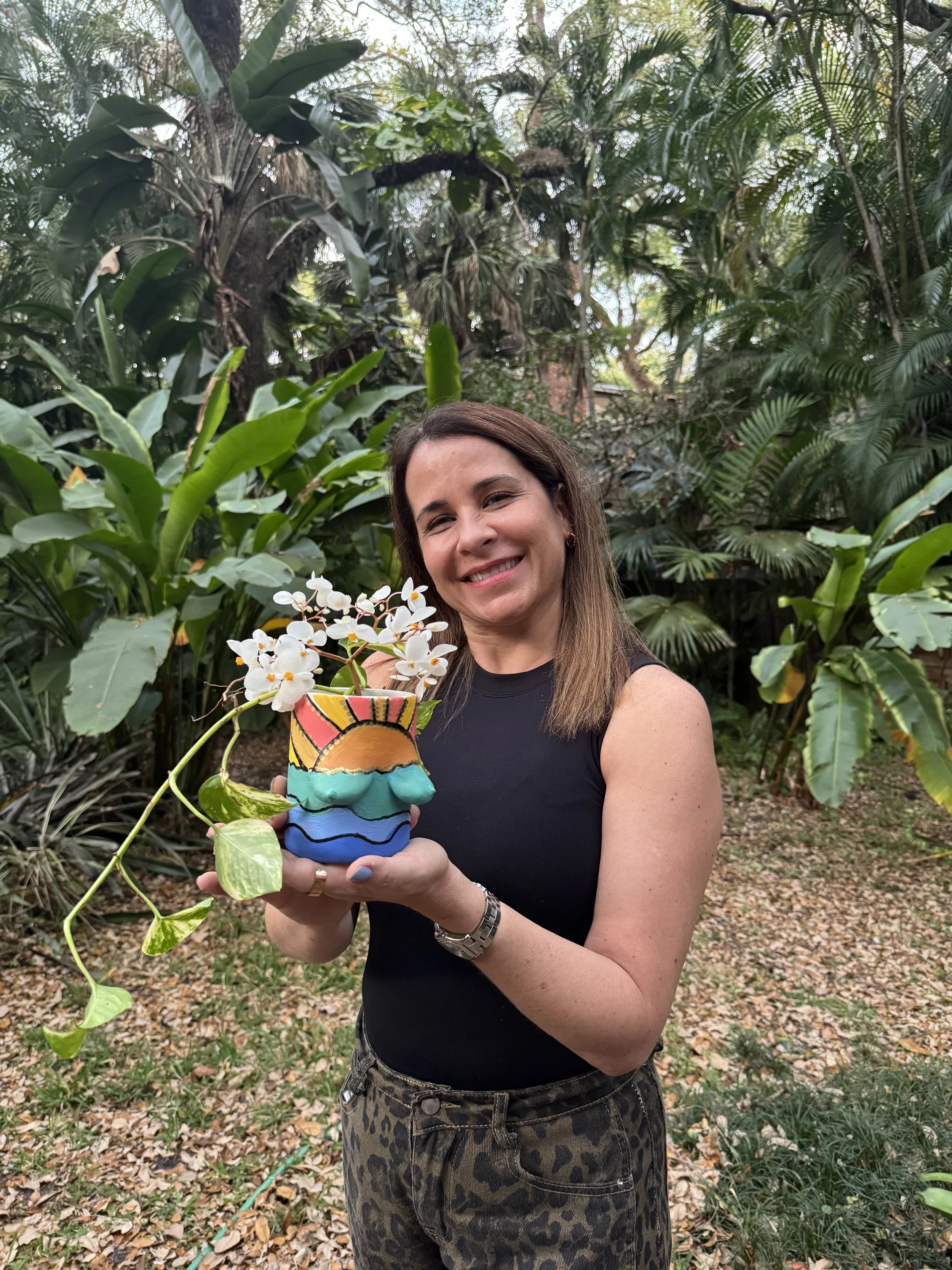 A woman with shoulder-length brown hair and a black sleeveless top, smiling and holding a colorful, painted flower pot with white flowers, standing outdoors with lush green tropical plants in the background.