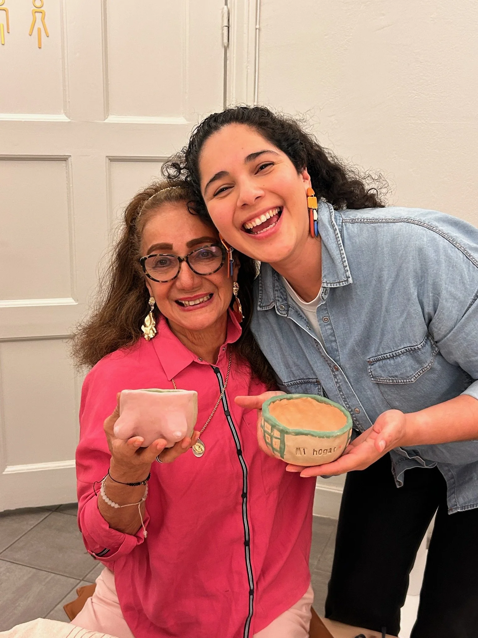 Two women, one older with glasses and earrings and the other younger, smiling and holding small ceramic pots, standing in front of a white door.