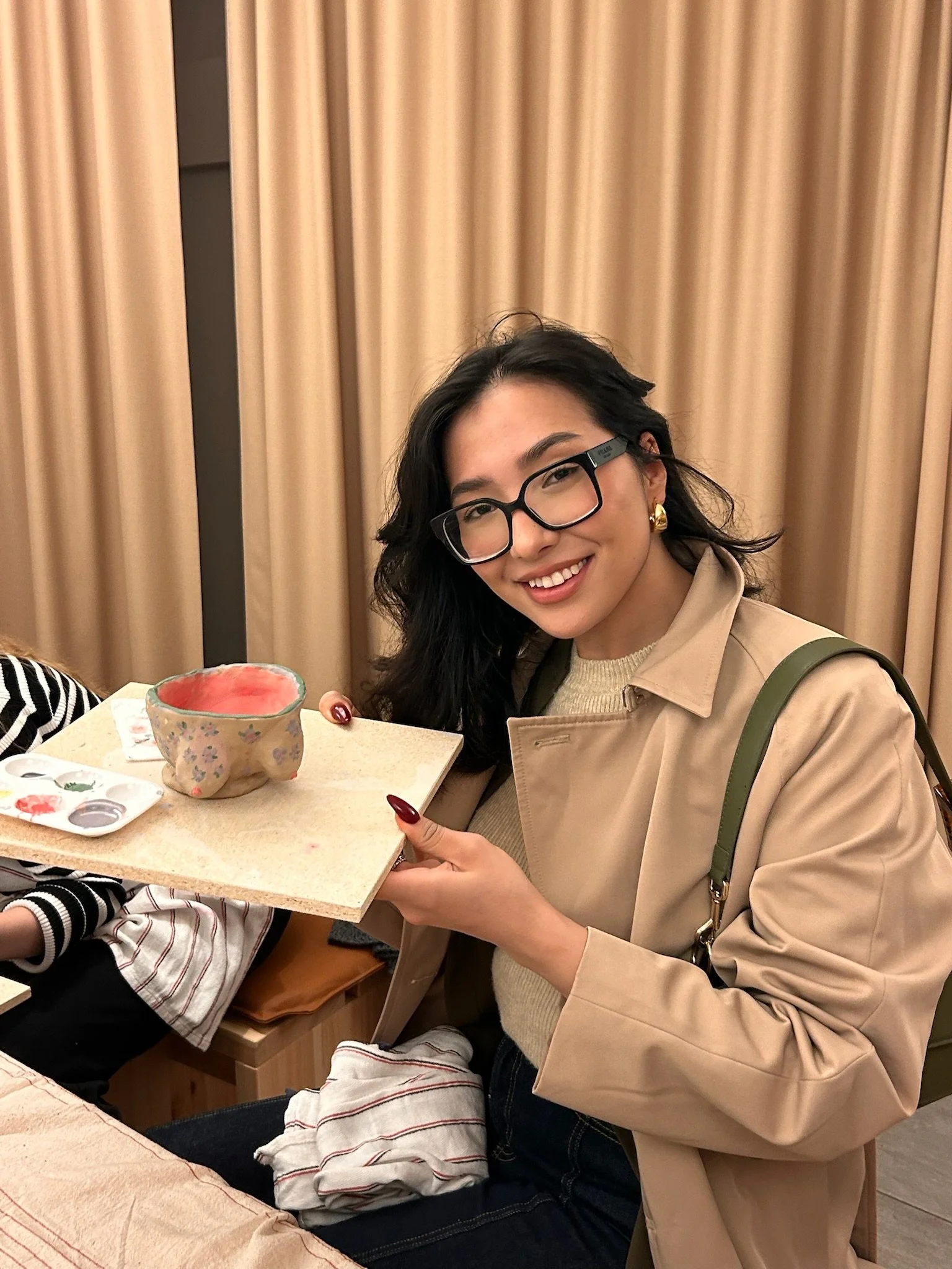 A woman with black hair, glasses, and gold earrings is smiling and holding a small ceramic bowl on a tray with paint and brushes.
