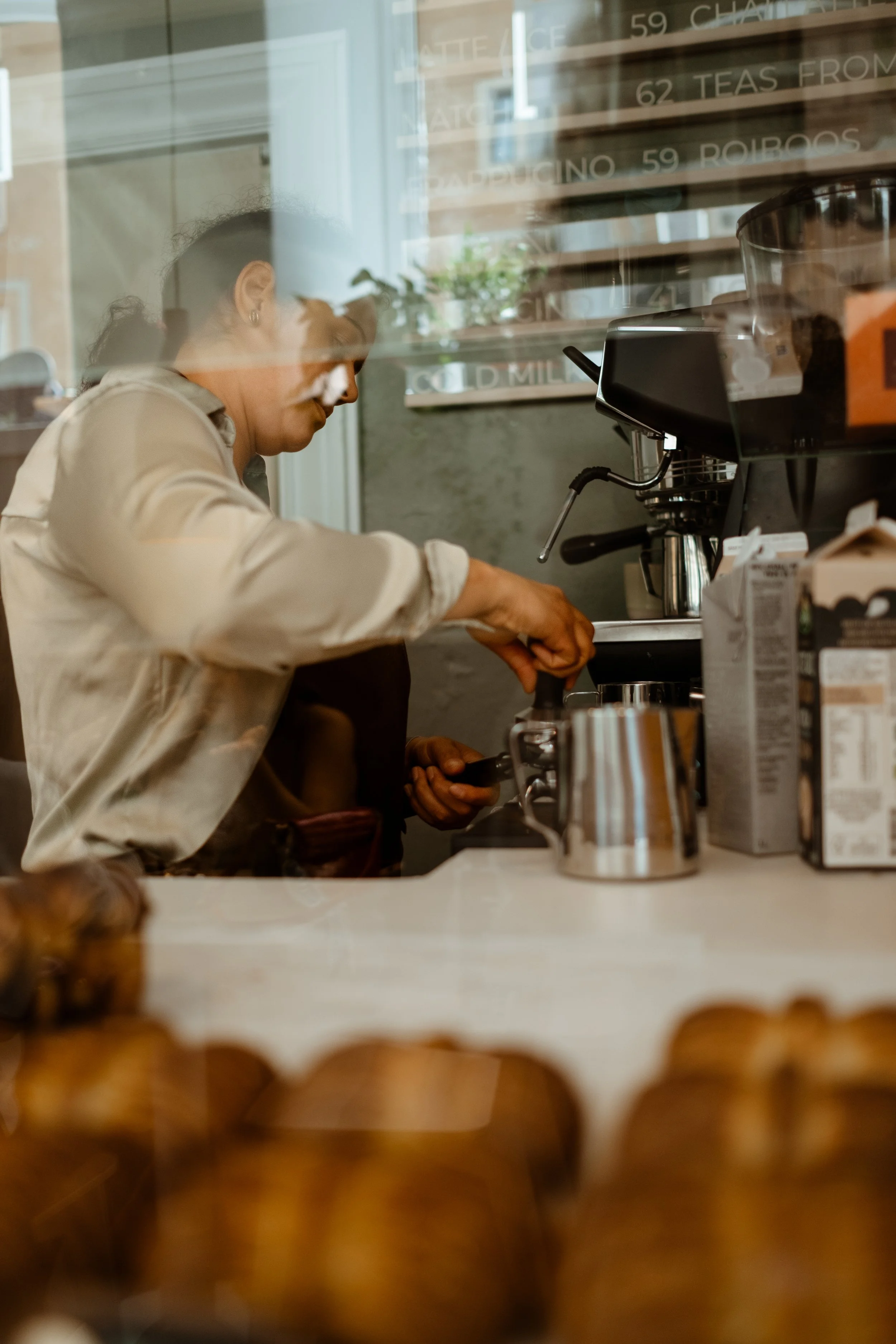 A woman making coffee behind a counter at a coffee shop, with baked goods in the foreground and a menu board behind her.