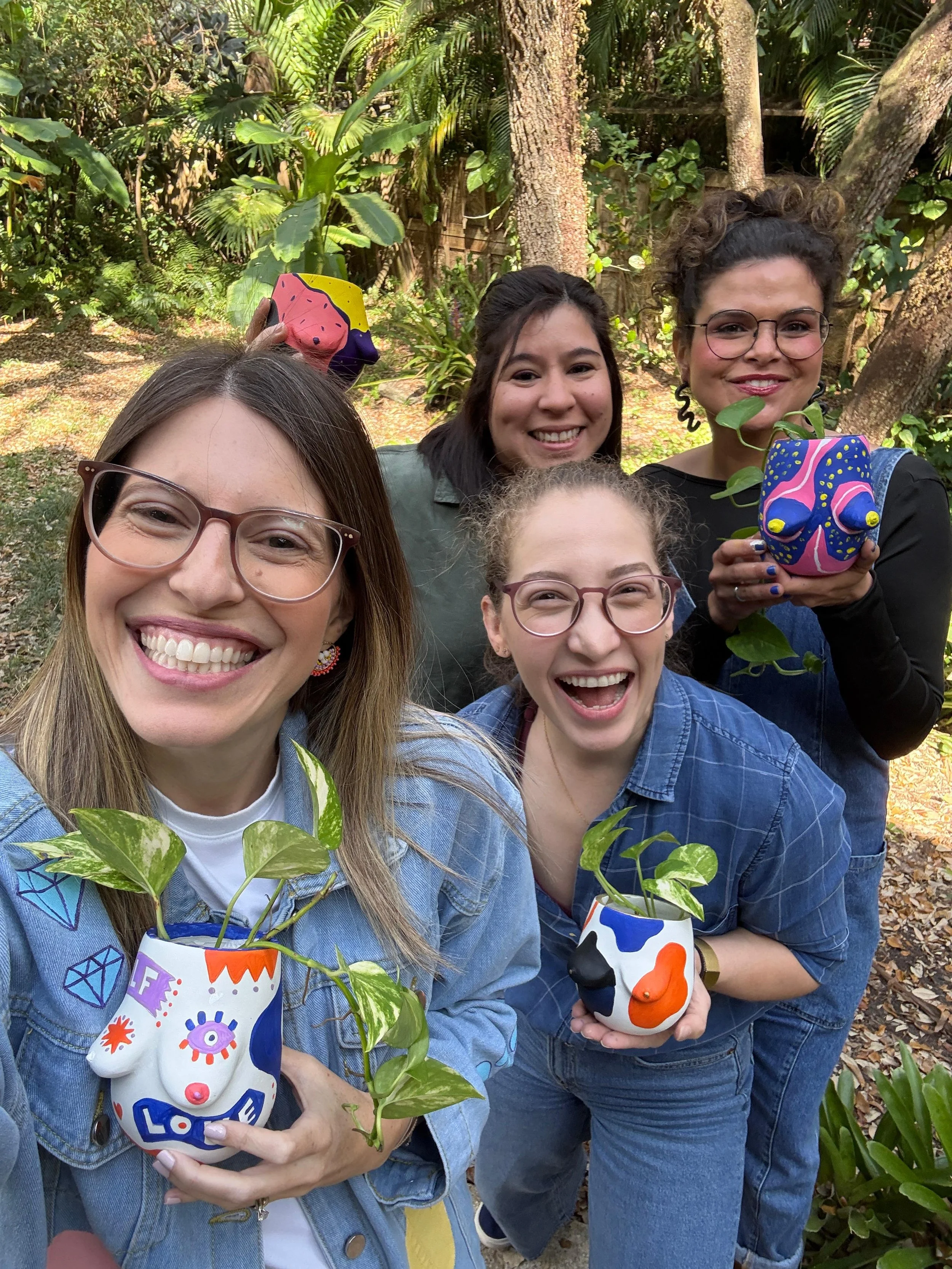 Group of five women smiling outdoors, holding colorful plant pots with green plants, amidst trees and lush greenery.