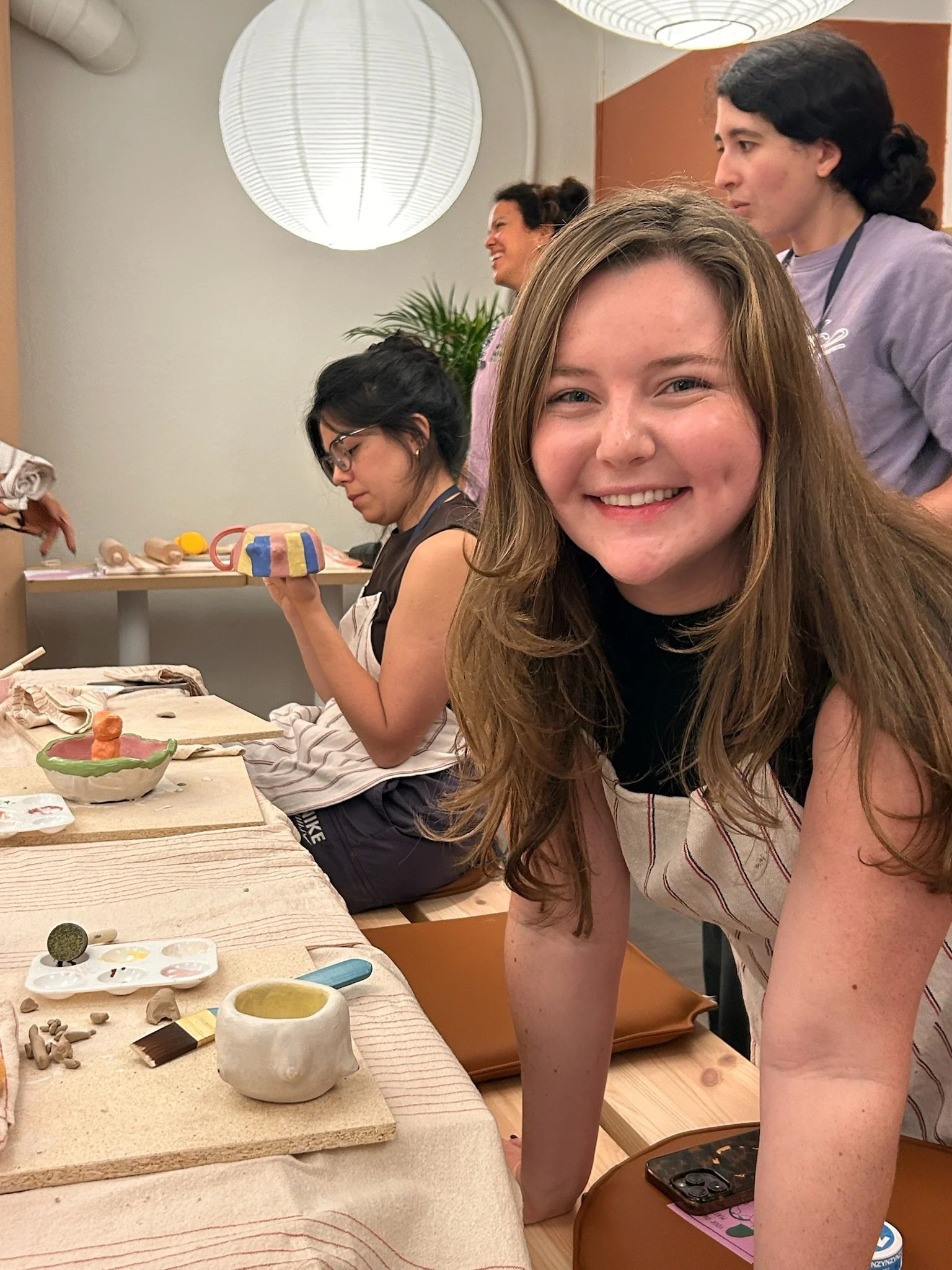 A group of women are participating in a pottery class, working on clay projects at a table with pottery tools and supplies.