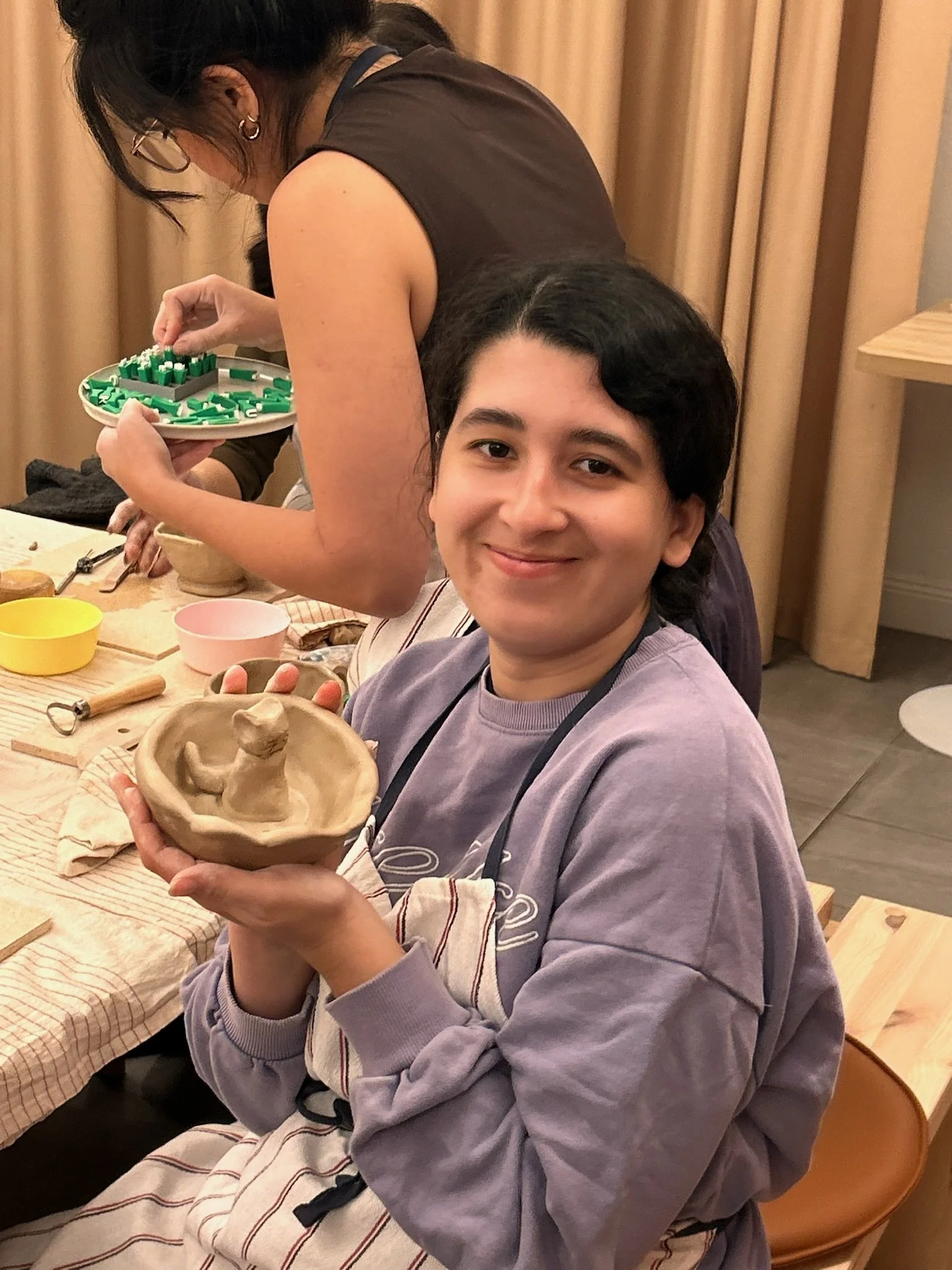 A person smiling and holding a small clay dish with a lizard-shaped sculpture inside in a pottery workshop.
