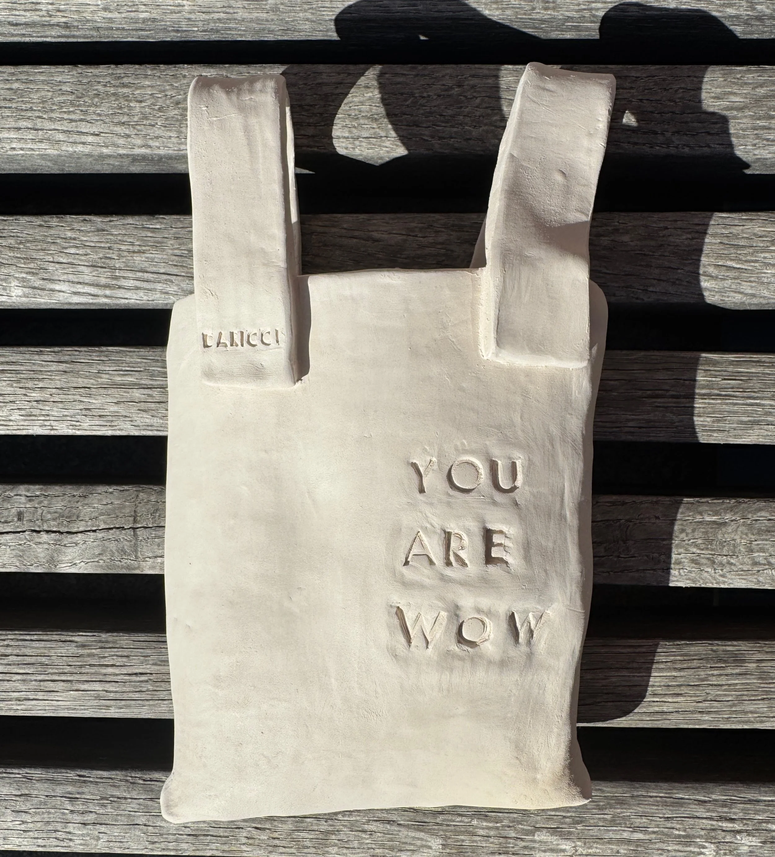 A white ceramic sculpture of a hand making a peace sign with the middle and index fingers raised, against a wooden background. The sculpture has the words 'LUCKY' on the thumb and 'YOU ARE WOW' on the palm.