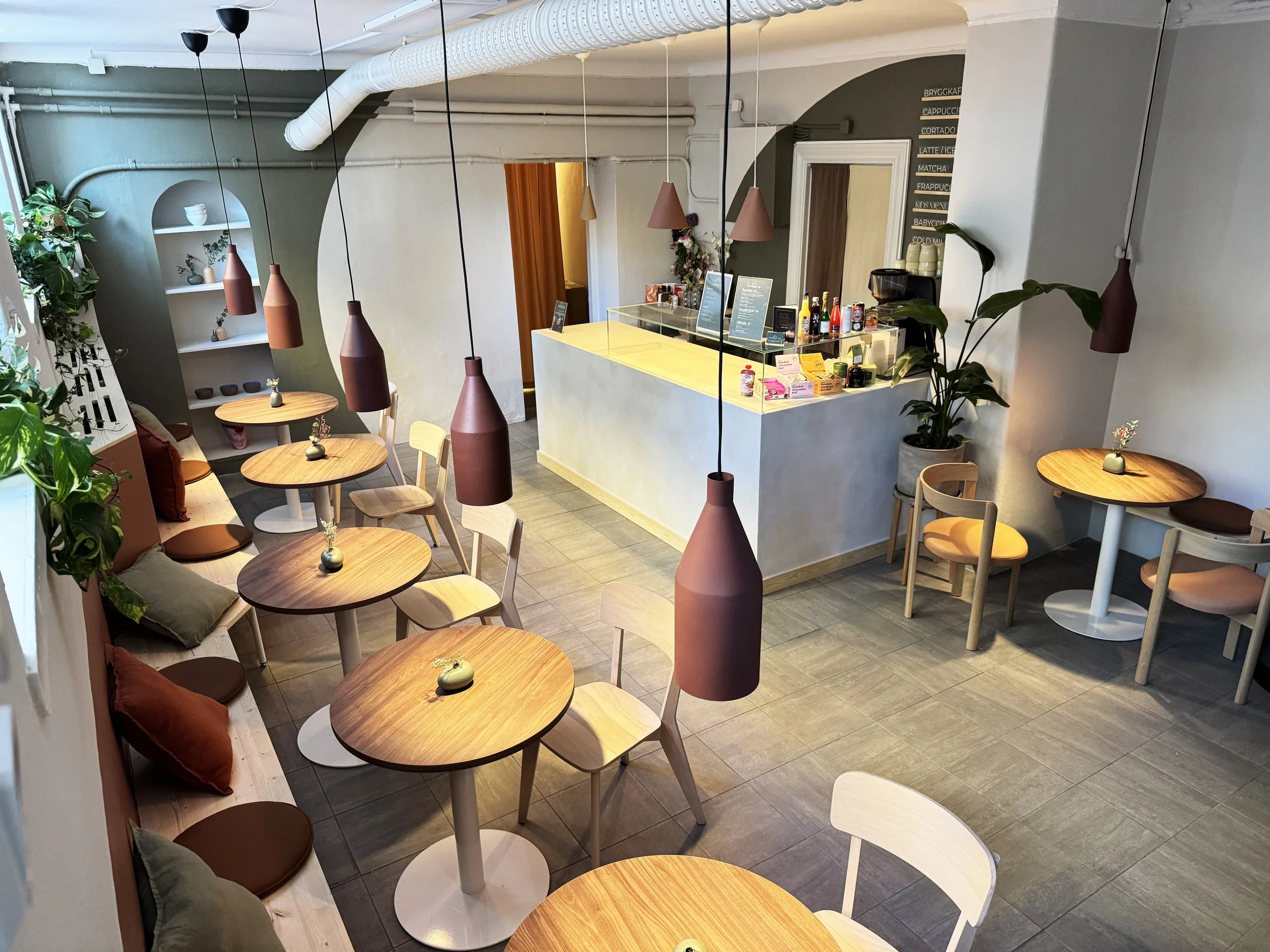 Interior of a cozy coffee shop with wooden tables, chairs, and pendant lights, decorated with plants and small vases on tables, and a counter with beverages and snacks.