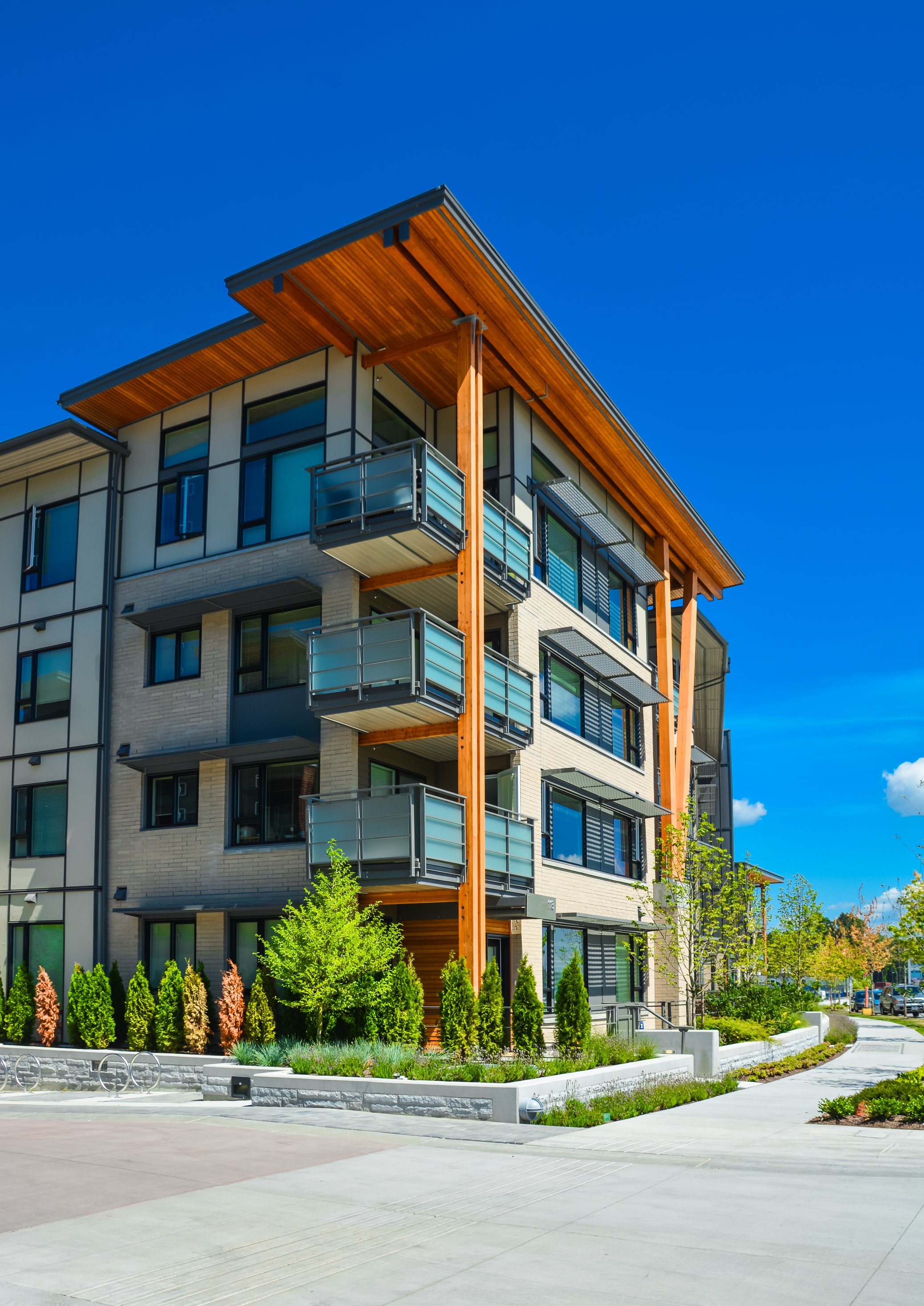 Modern multi-story apartment building with balconies, surrounded by green trees and a landscaped area, under a clear blue sky.