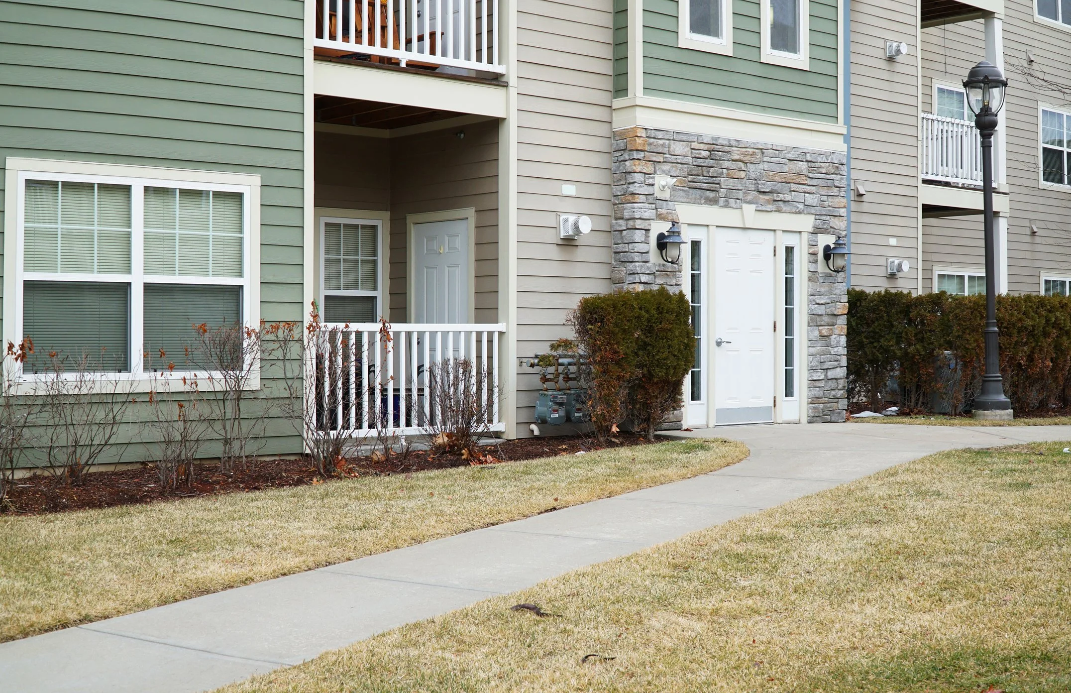 Exterior view of a modern apartment building with beige siding, stone accents, white window frames, and a small balcony. A sidewalk and grassy area with bushes are in the foreground, and a streetlamp is on the right side.