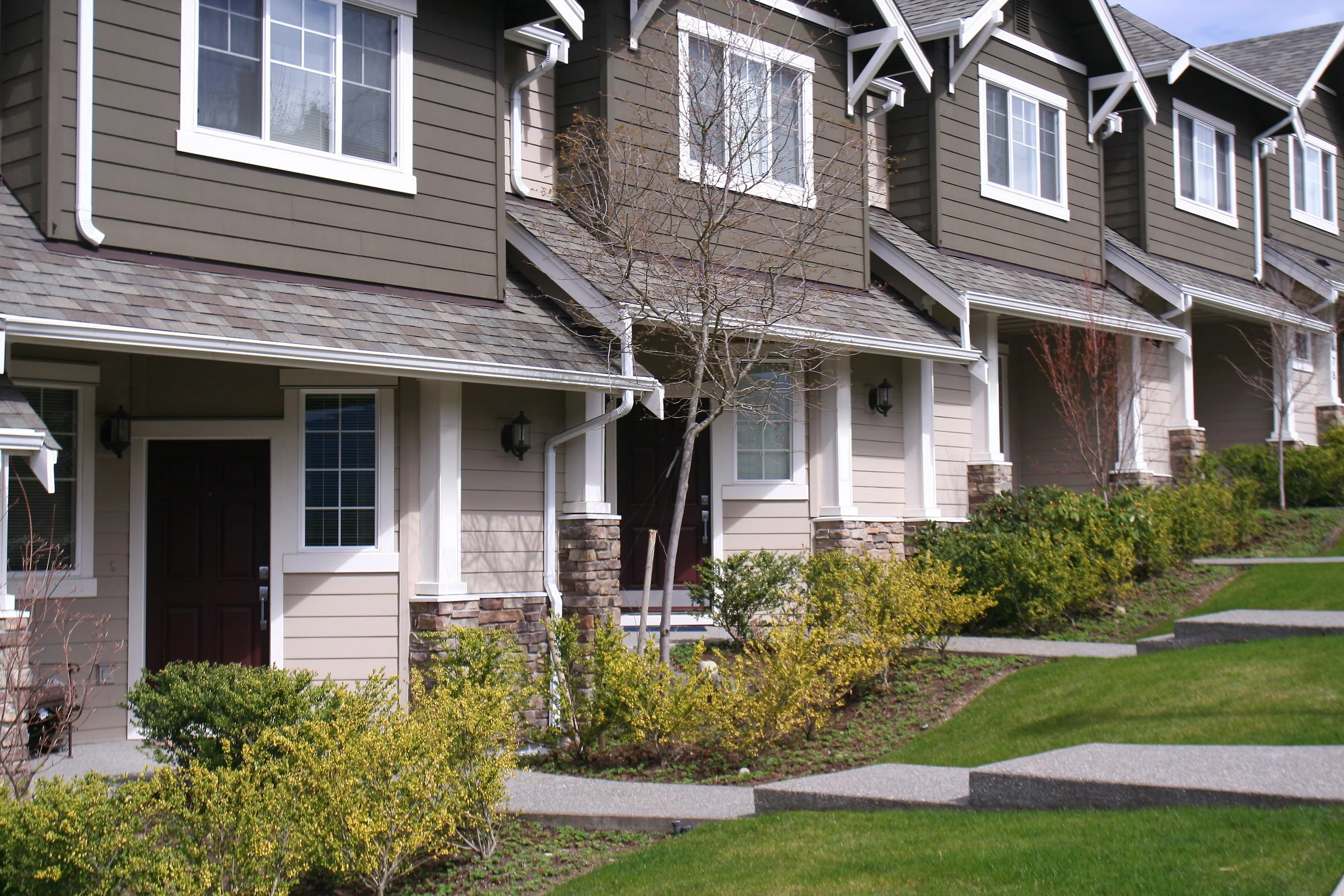 Row of beige townhouses with brown roofs, white trim, and decorative gables. Small front yard with plants, bushes, and a sidewalk.