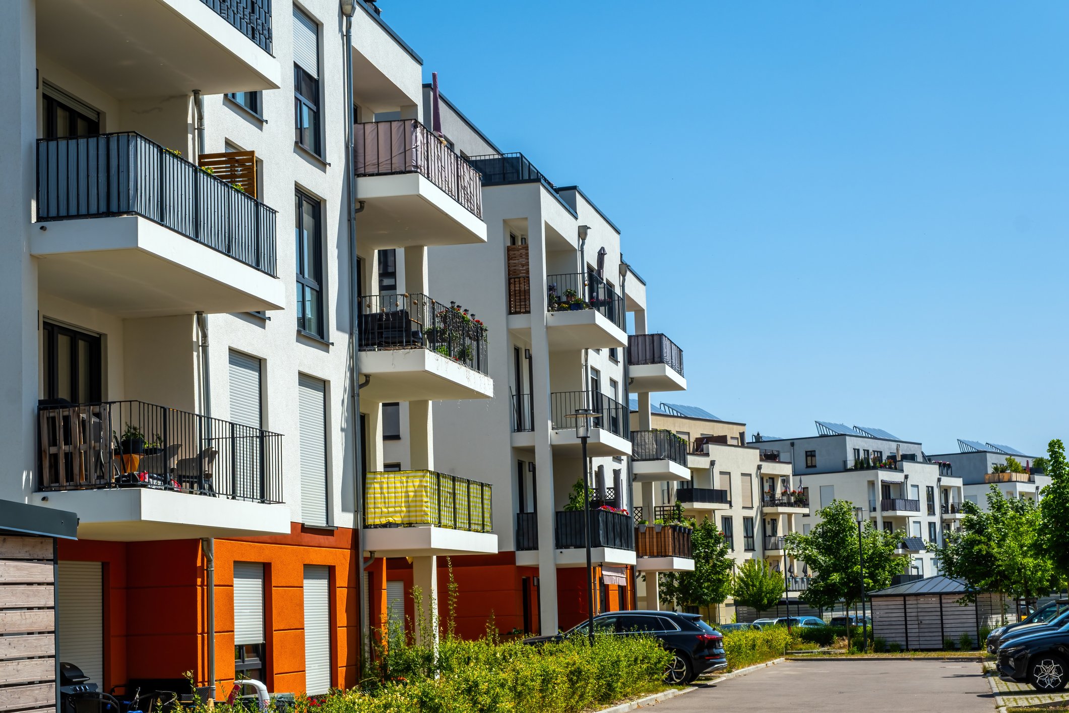 Modern white residential apartment buildings with balconies, some decorated with plants and outdoor furniture, set against a clear blue sky, with parked cars and a sidewalk with trees in the foreground.