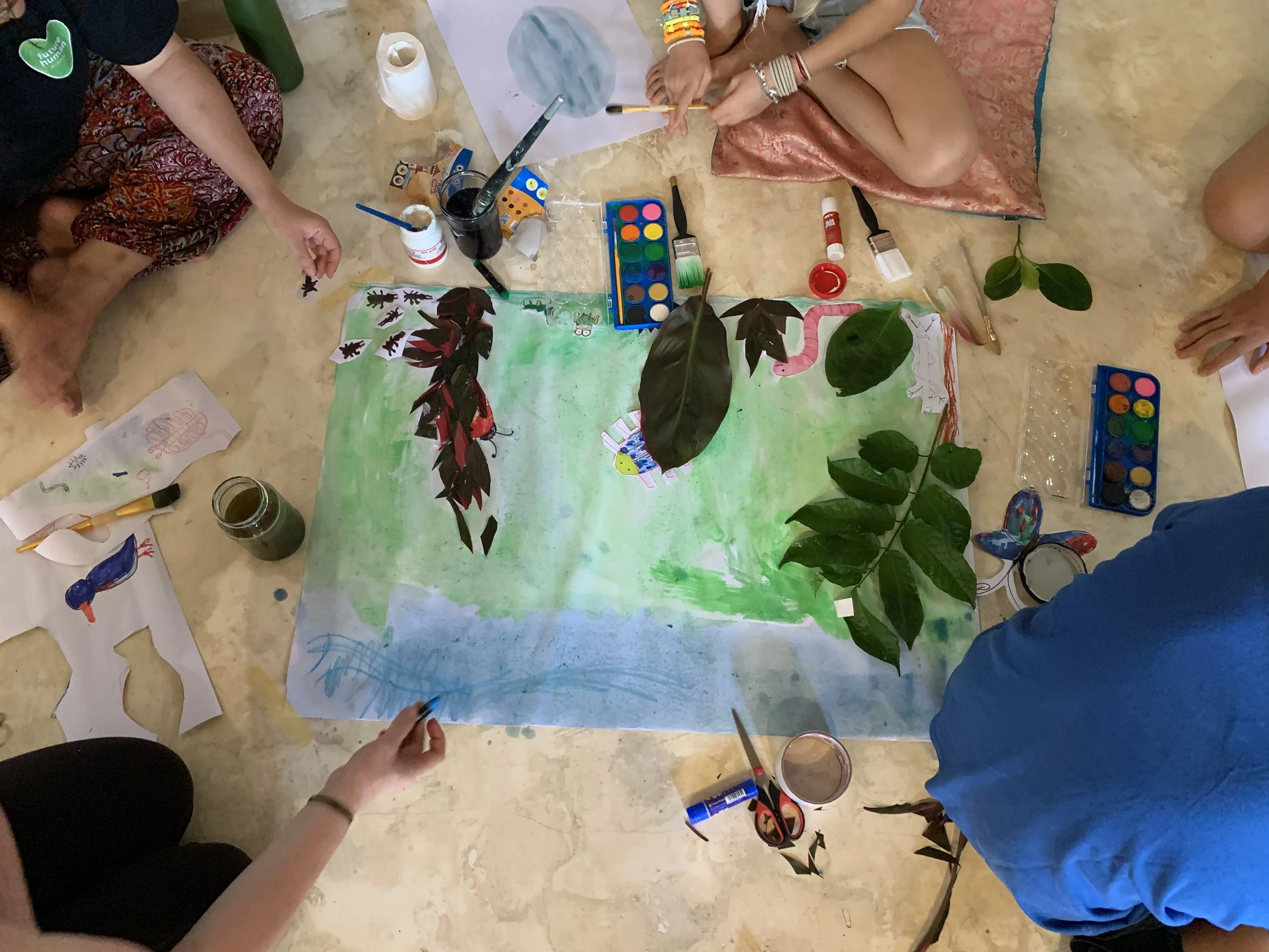 People creating a nature-themed painting on a large sheet of paper, surrounded by leaves, paints, brushes, and drawing papers.