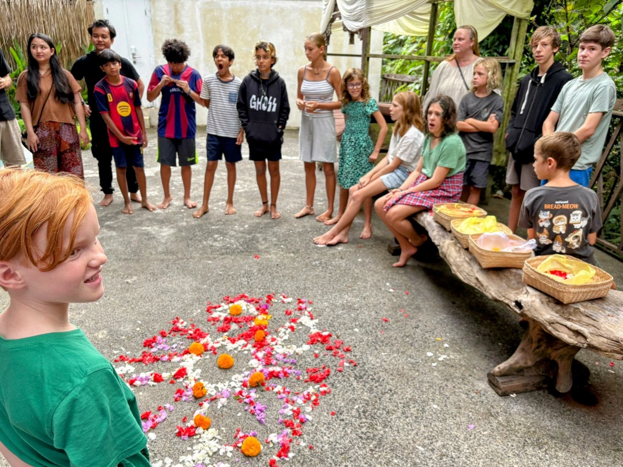 Children and adults gathered outdoors around a floral arrangement on the ground, with some sitting on a bench and others standing, during a cultural or recreational event.