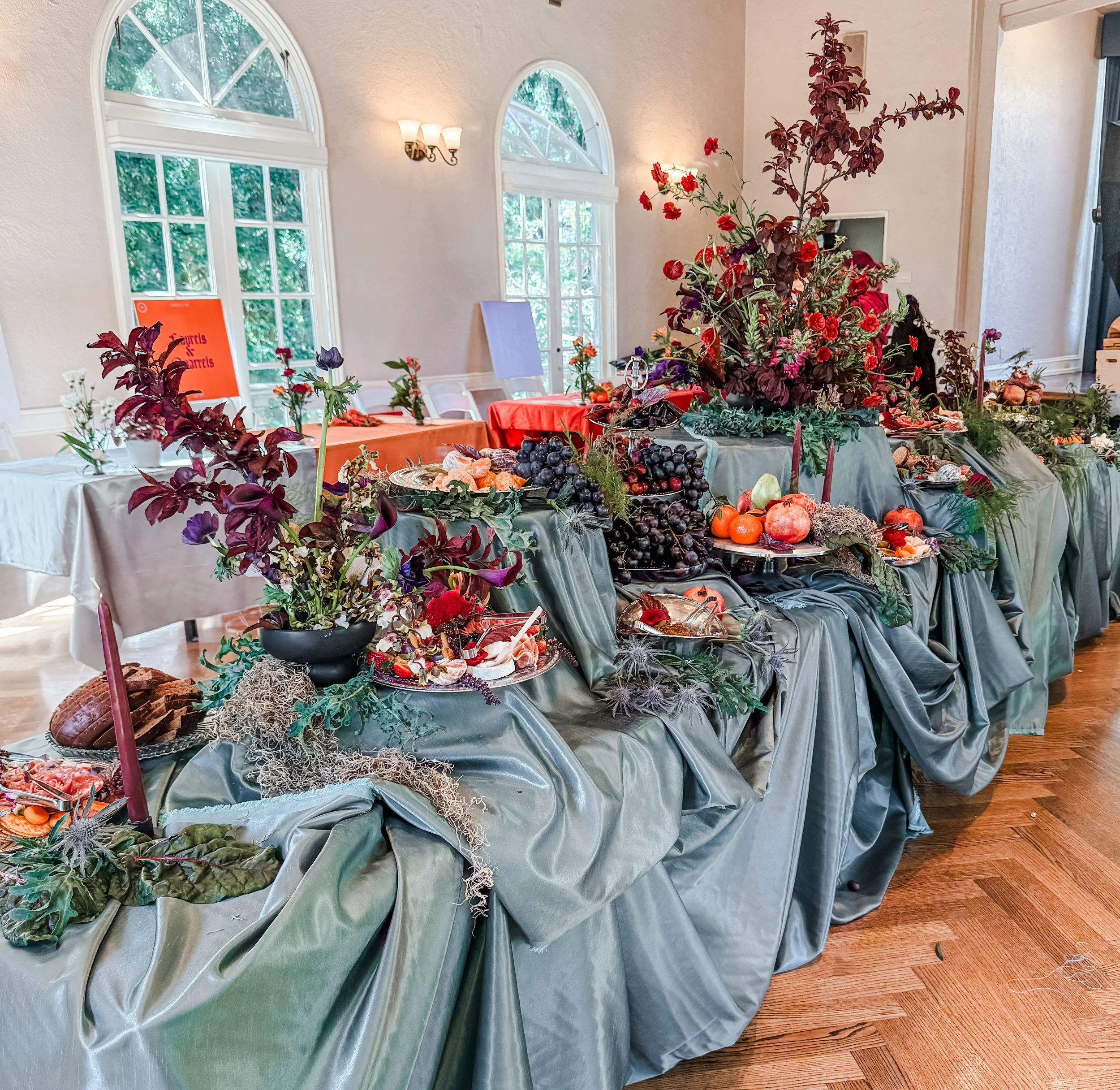 Elegant banquet table with dark green satin tablecloth and fall-themed decorations, including grapes, pumpkins, flowers, and candles, set in a bright room with large arched windows.