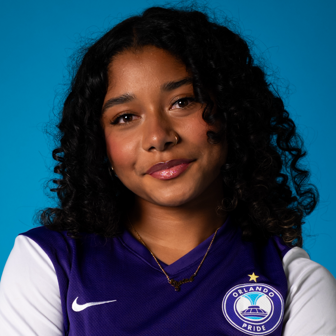 A young woman with curly black hair, wearing a purple Orlando Pride soccer jersey, with a gold necklace and a nose piercing, smiling against a blue background.