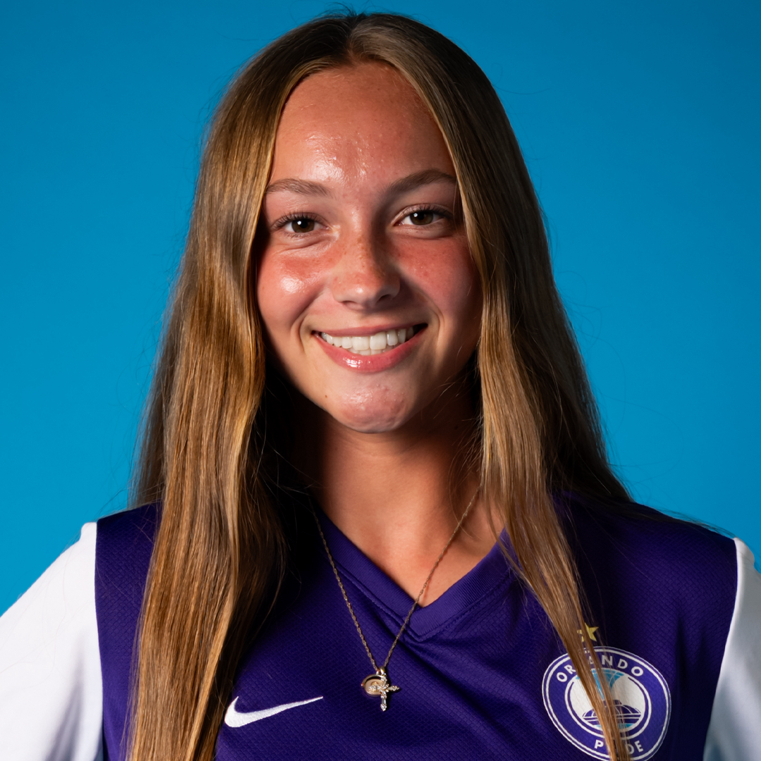 Young woman with long blonde hair smiling, wearing an Orlando Pride soccer jersey and a necklace with a cross pendant, against a blue background.