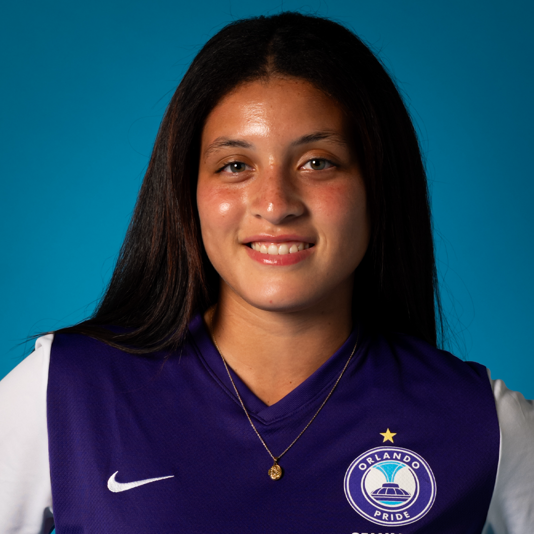 Young woman with long dark hair smiling, wearing a purple Orlando Pride soccer jersey with white sleeves, a gold star above the team logo, and a gold necklace, against a blue background.