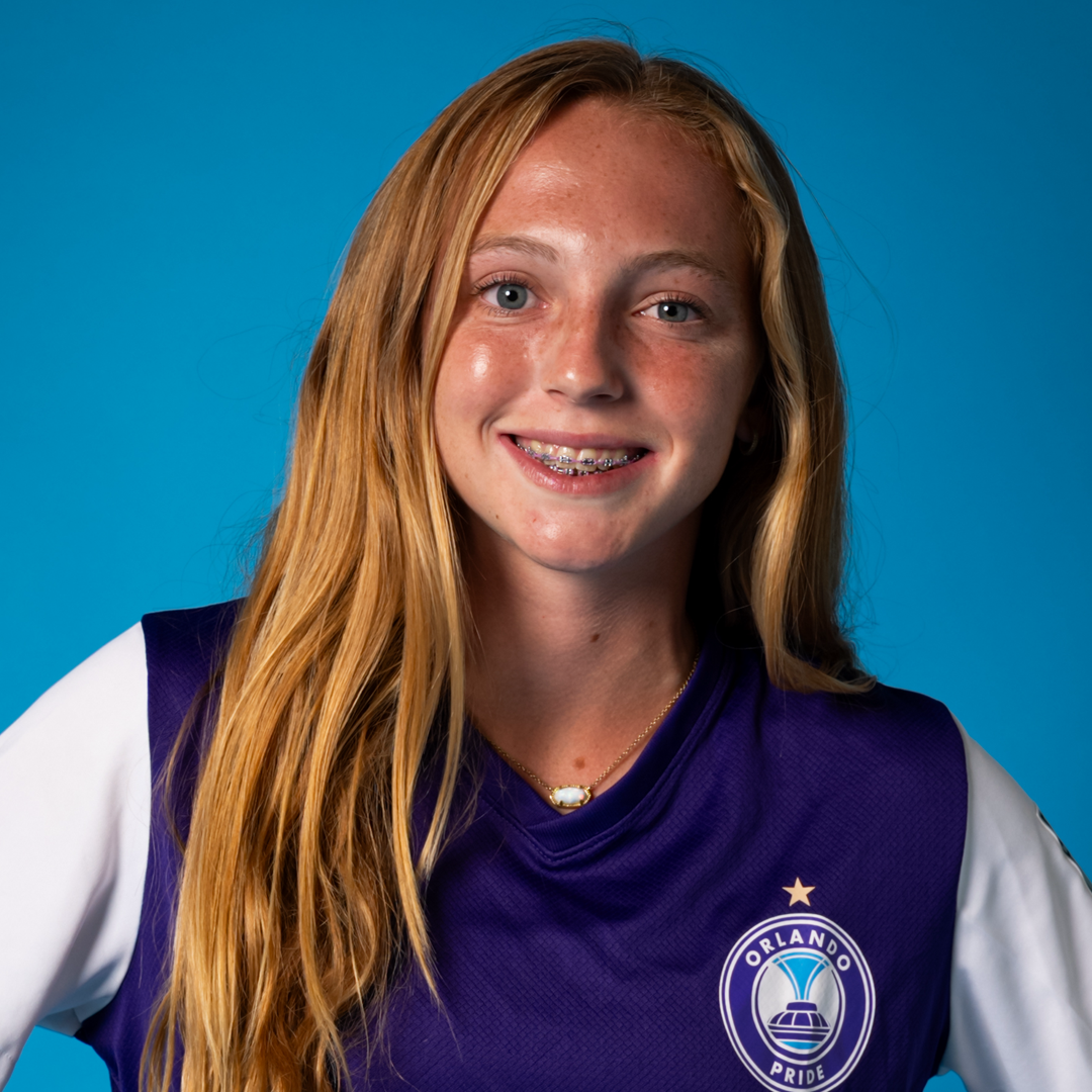 A teenage girl with long red hair smiling, wearing braces, a purple and white sports jersey with an Orlando Pride logo, and a small necklace, against a blue background.