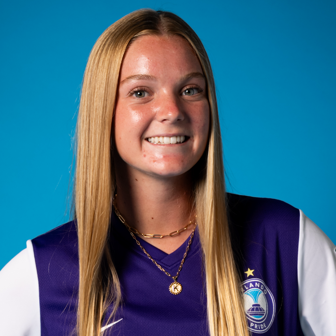 Young woman with long blonde hair wearing an Orlando Pride soccer jersey and layered gold necklaces, smiling against a blue background.