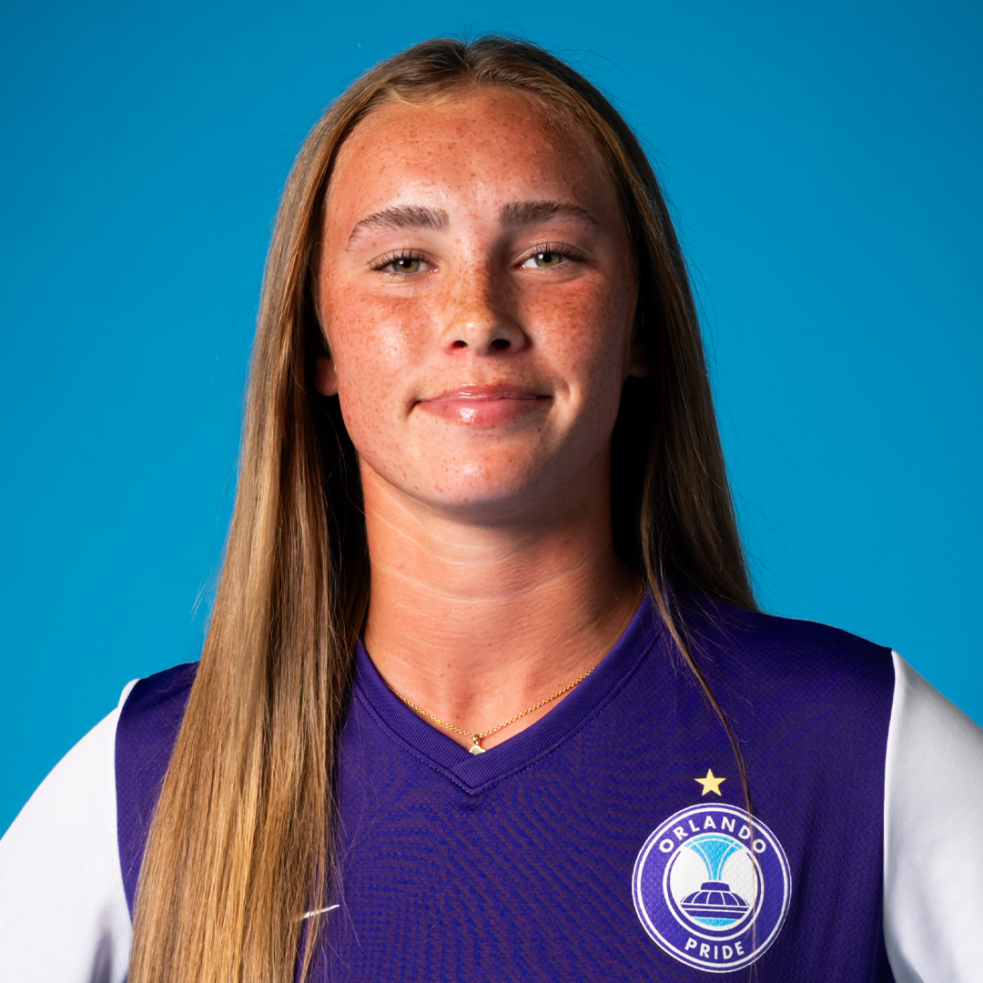 Portrait of a young woman with long brown hair, wearing an Orlando Pride soccer jersey, against a blue background.