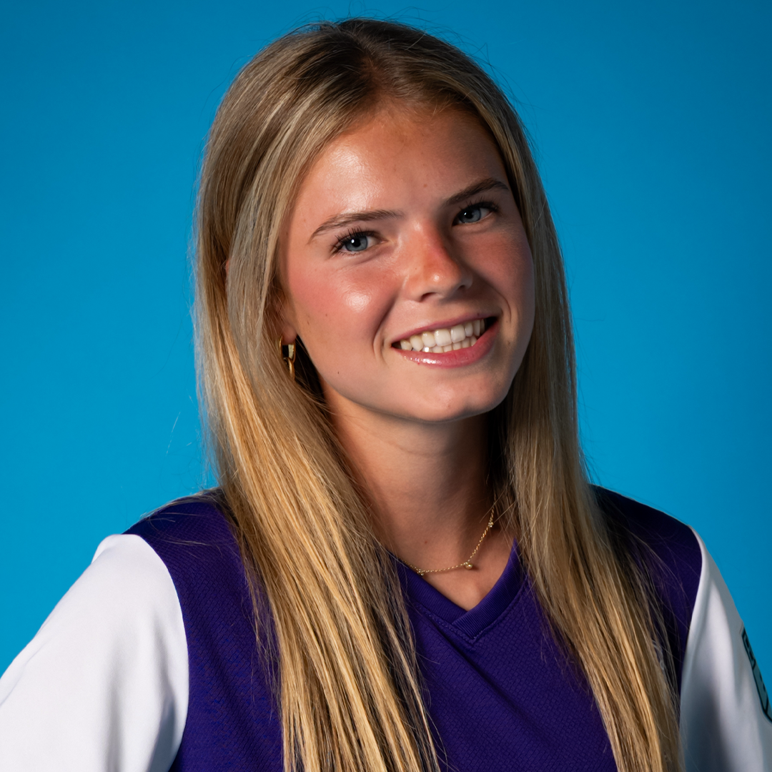 A young woman with long blonde hair, blue eyes, and light skin, smiling at the camera, wearing a purple sports shirt with white sleeves, gold earrings, and a gold necklace, against a blue background.