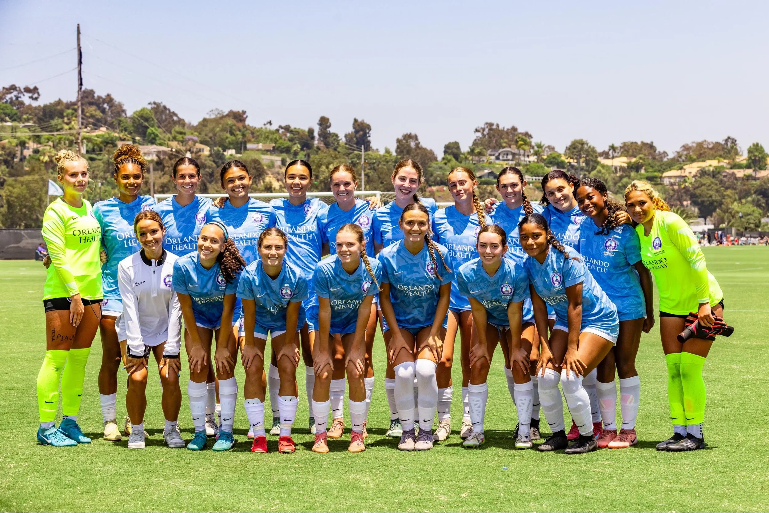 A women's soccer team posing on a grass field, wearing blue uniforms with Orlando Health logo, with two goalkeepers in neon green uniforms, on a sunny day with hills and houses in the background.