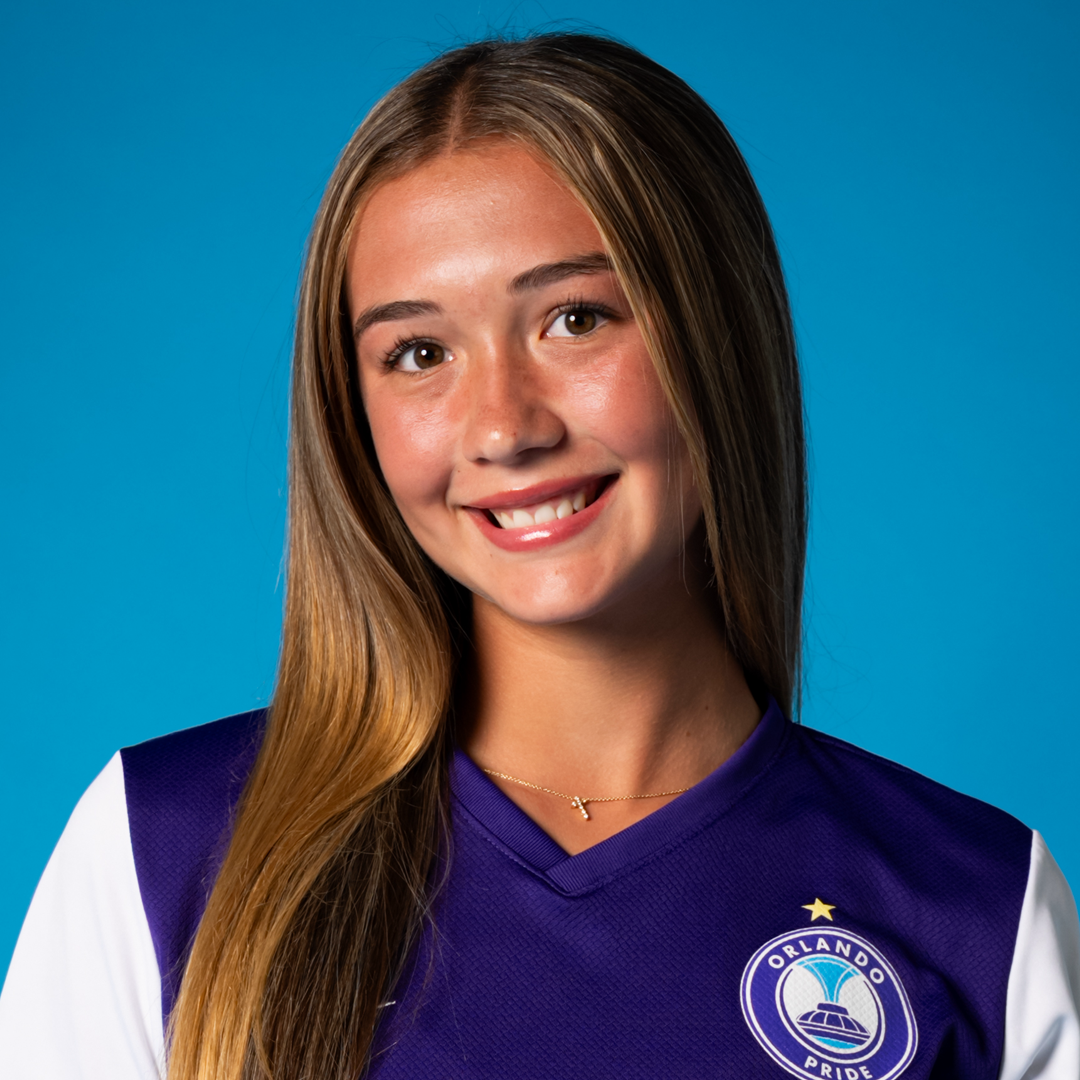 Portrait of a young woman with long light brown hair, smiling, wearing a purple Orlando Pride soccer jersey with a logo, against a blue background.