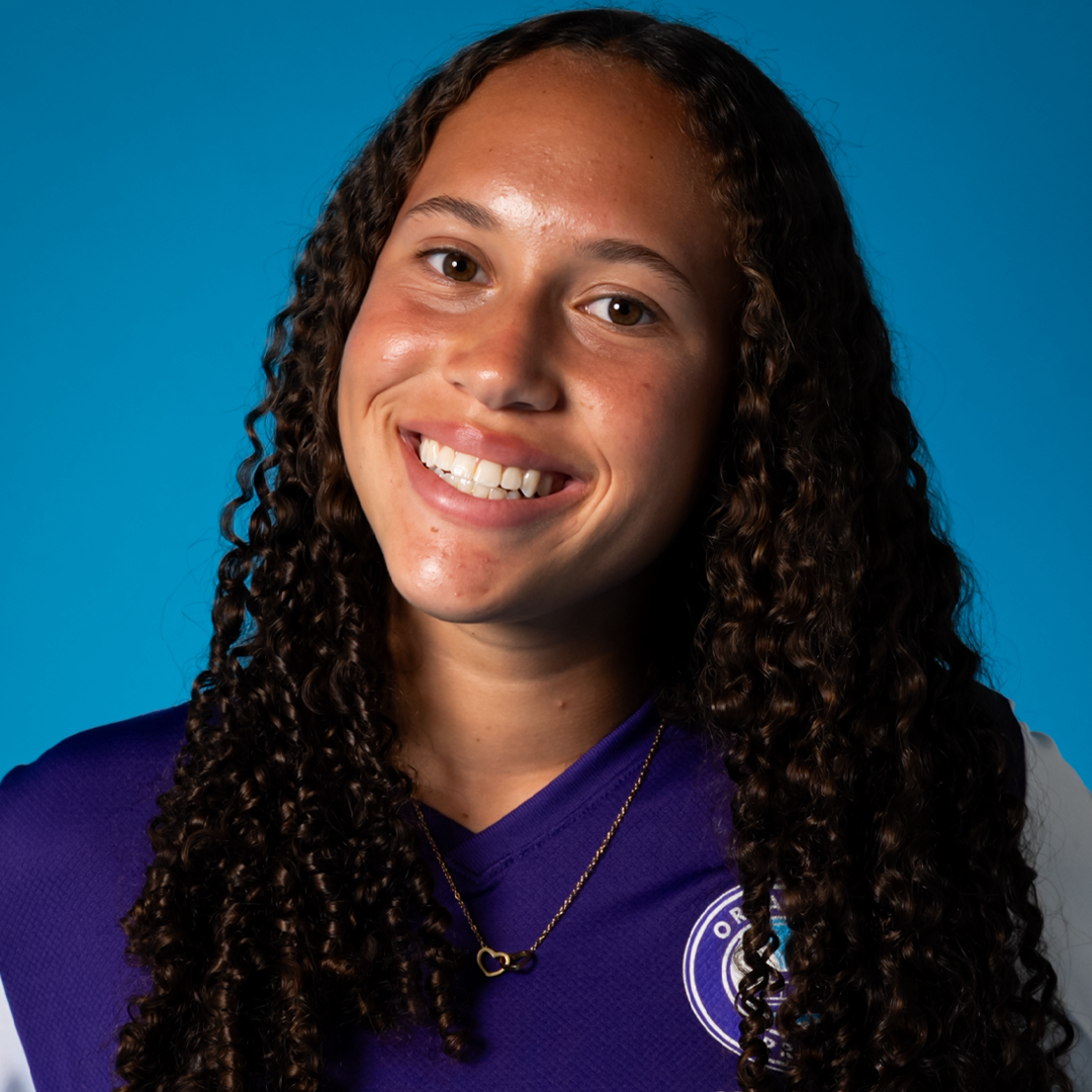 A young woman with long, curly hair, smiling and showing her teeth, wearing a purple sports jersey and a gold necklace with a heart-shaped pendant, against a blue background.
