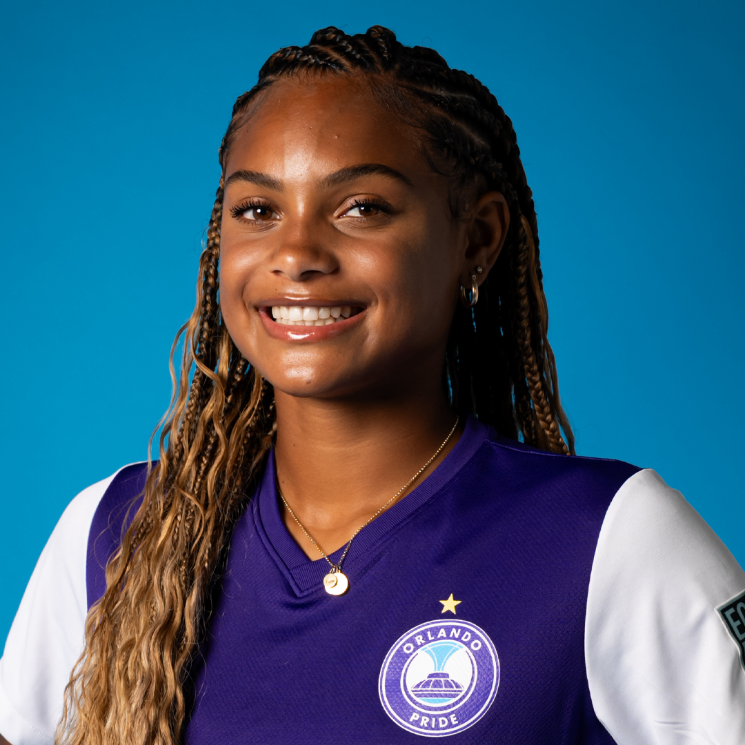 A young woman with long braided hair smiling, wearing a purple and white Orlando Pride soccer jersey, gold necklace, and earrings, against a blue background.