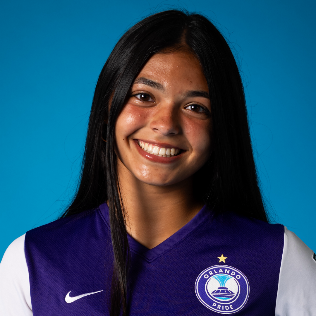 Young woman with long black hair smiling, wearing an Orlando Pride soccer jersey, standing against a blue background.