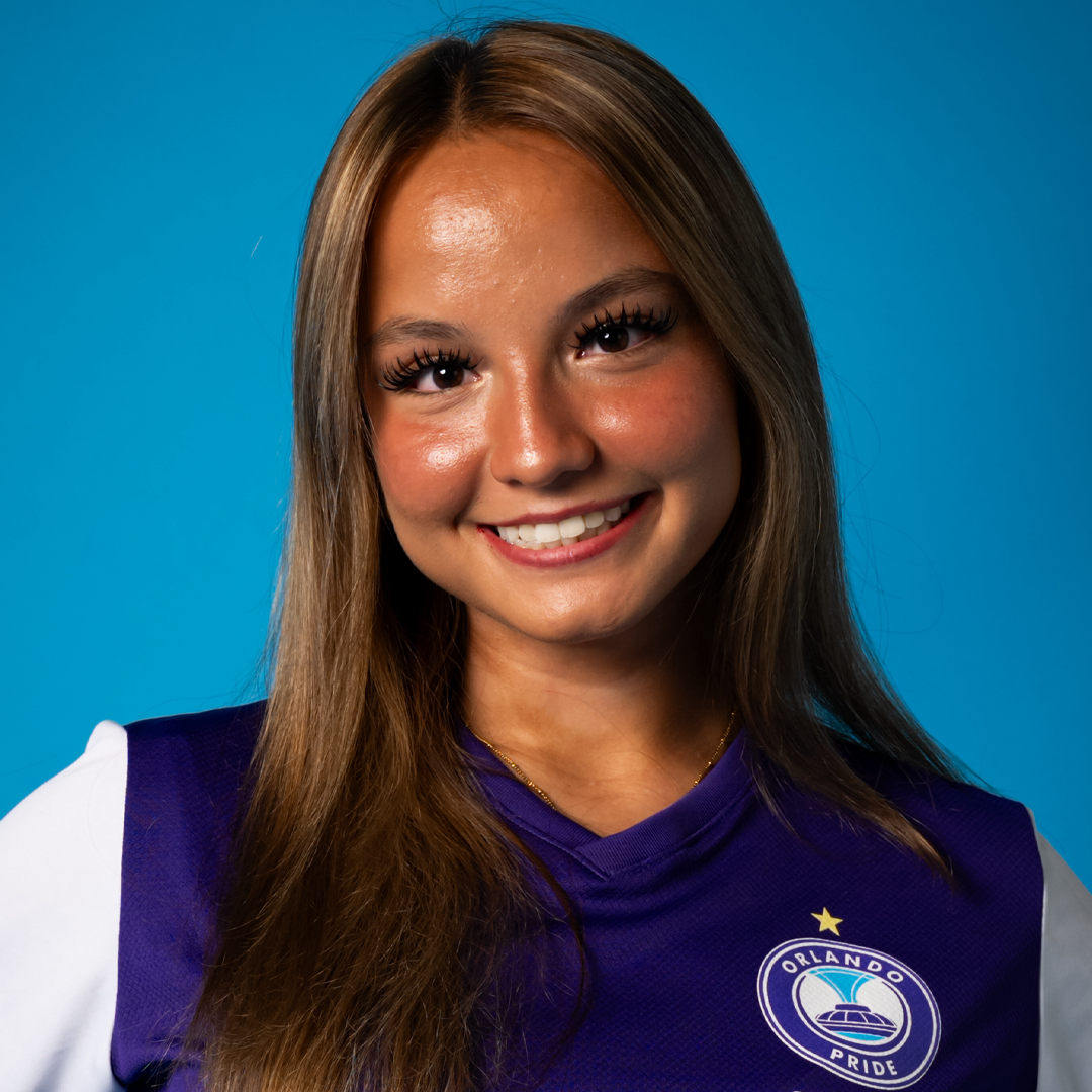 Smiling young woman with long brown hair wearing a purple Orlando Pride soccer jersey against a blue background.