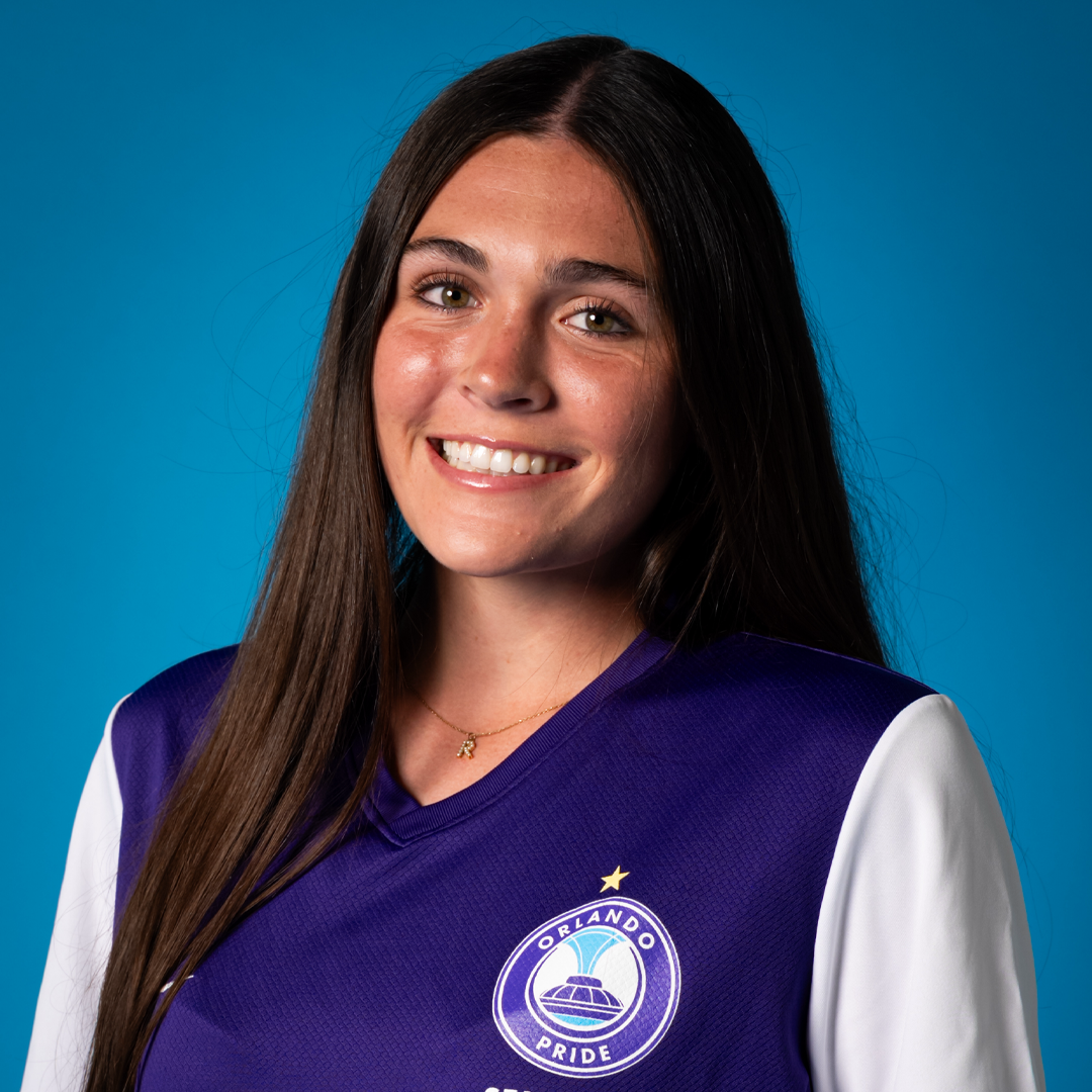 A woman with long brown hair wearing a purple and white Orlando Pride soccer jersey, smiling in front of a blue background.