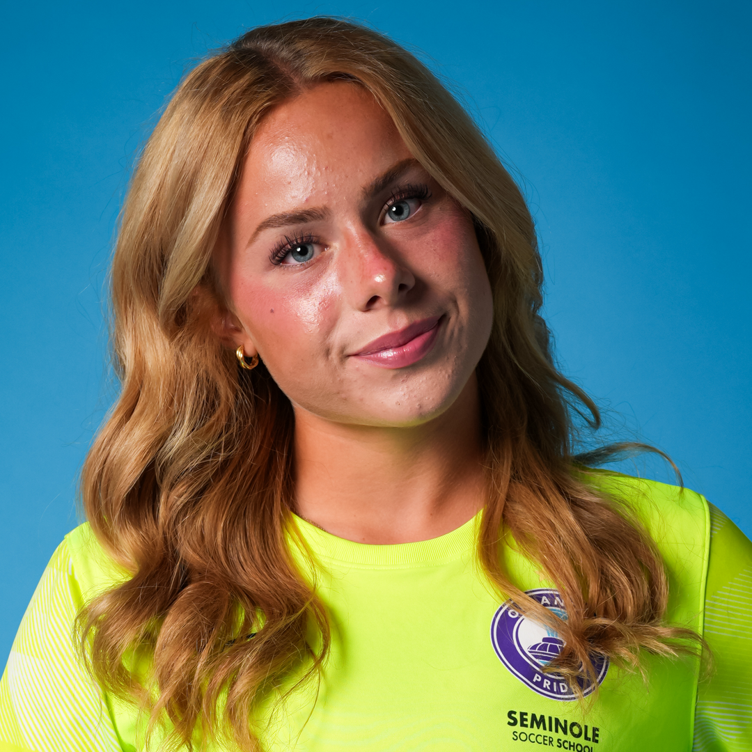Portrait of a young woman with long wavy red hair, wearing a neon yellow Seminole Soccer School shirt, against a light blue background.