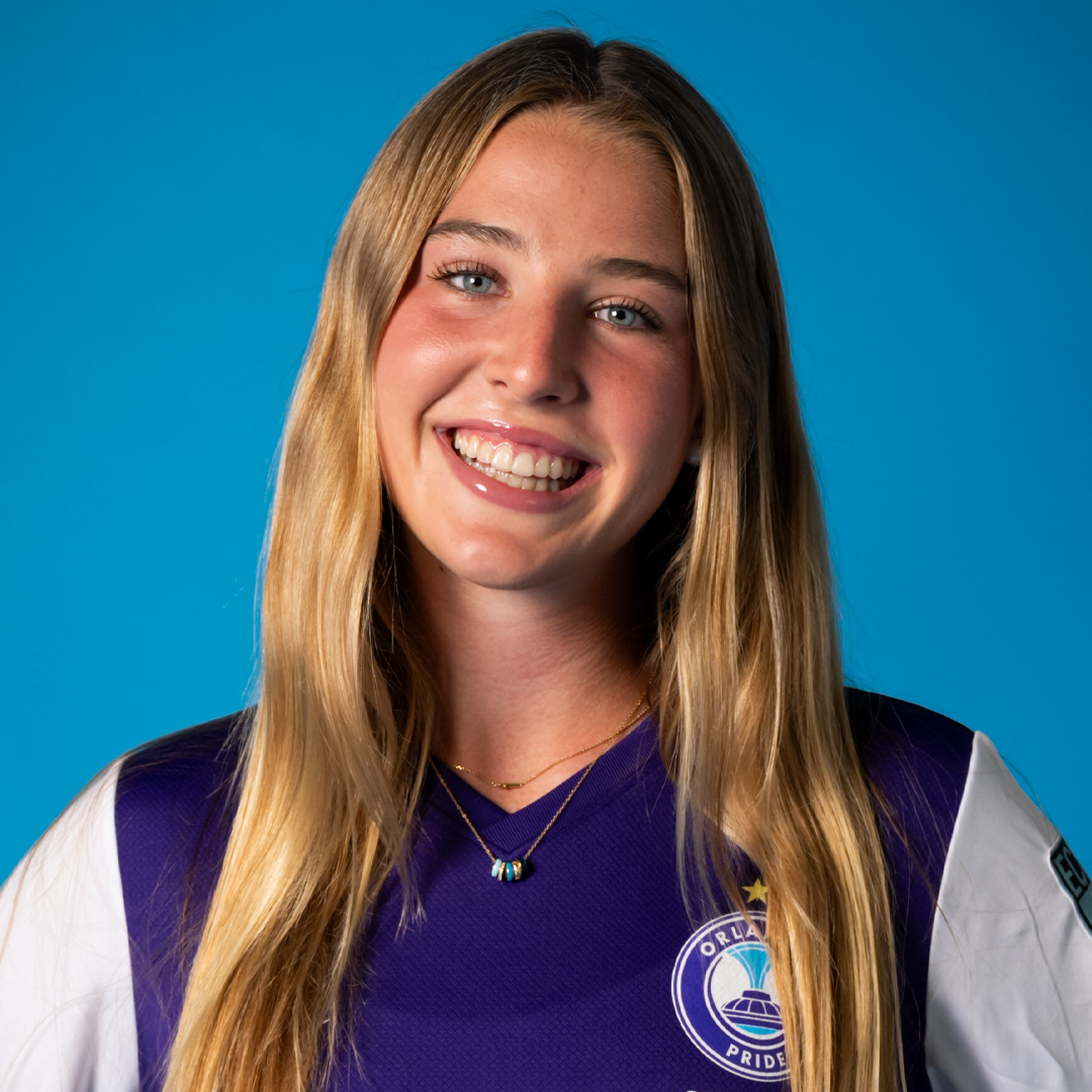 Young woman with blonde hair smiling, wearing a purple sports jersey with a logo and a necklace, against a blue background.