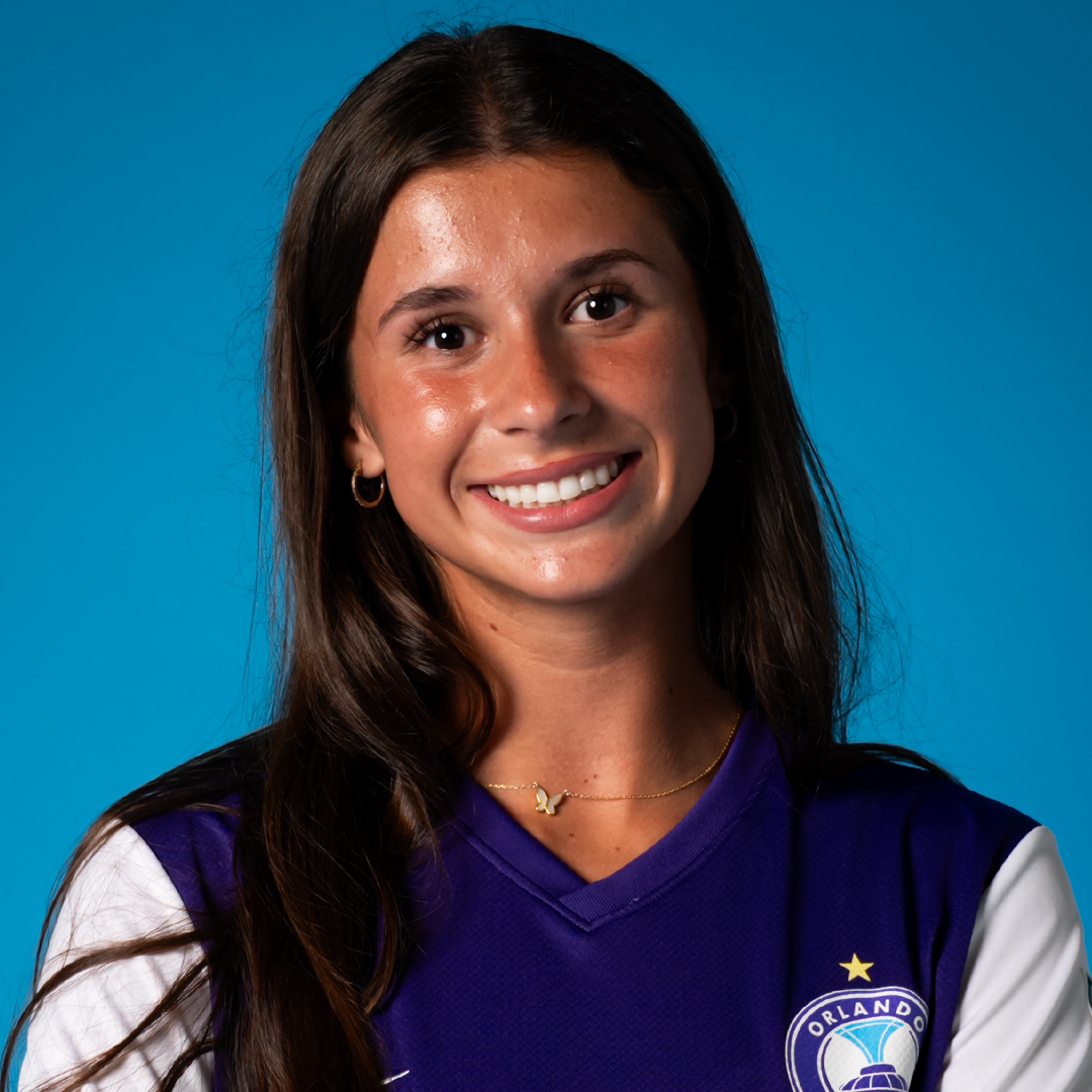 Young woman with long brown hair smiling, wearing a purple Orlando soccer jersey and gold jewelry, against a blue background.