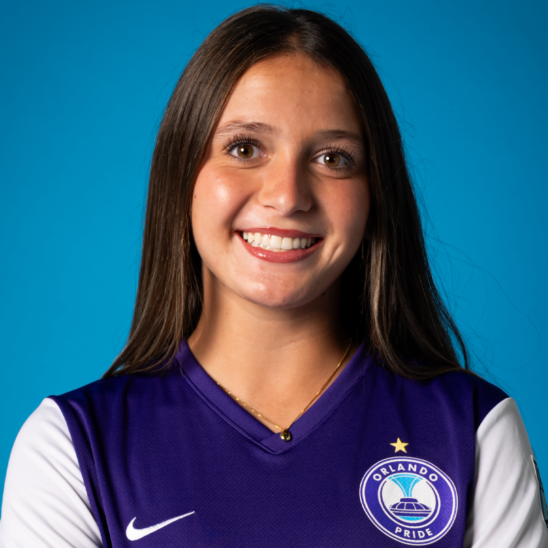 A young woman smiling, wearing a purple Orlando Pride soccer jersey with white sleeves and a Nike logo, against a blue background.