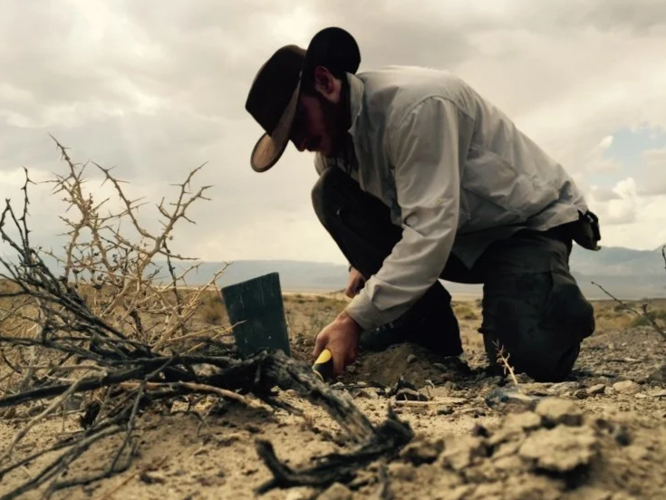 A man wearing a hat, sunglasses, and a light-colored jacket kneeling on dry, cracked ground in an arid landscape, examining or working with a small piece of equipment near a dry, leafless branch.