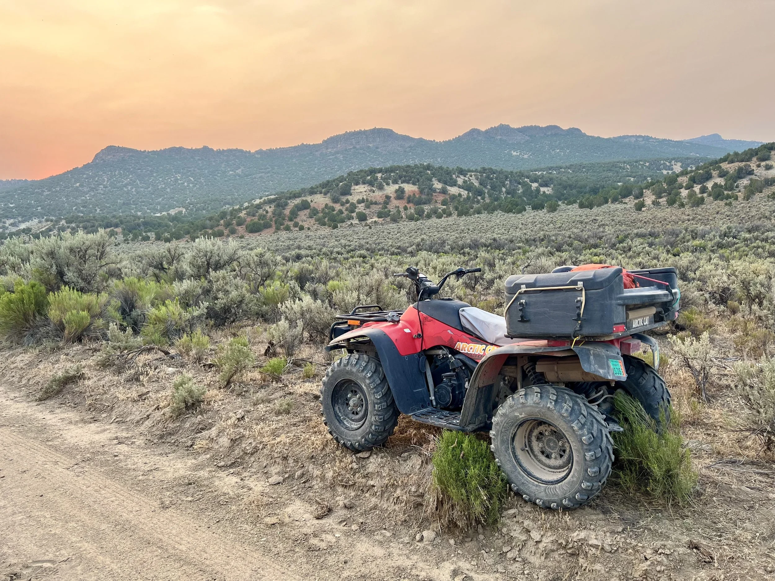 A dirt bike with large tires and a storage box parked on a dirt trail in a mountainous desert landscape at sunset.