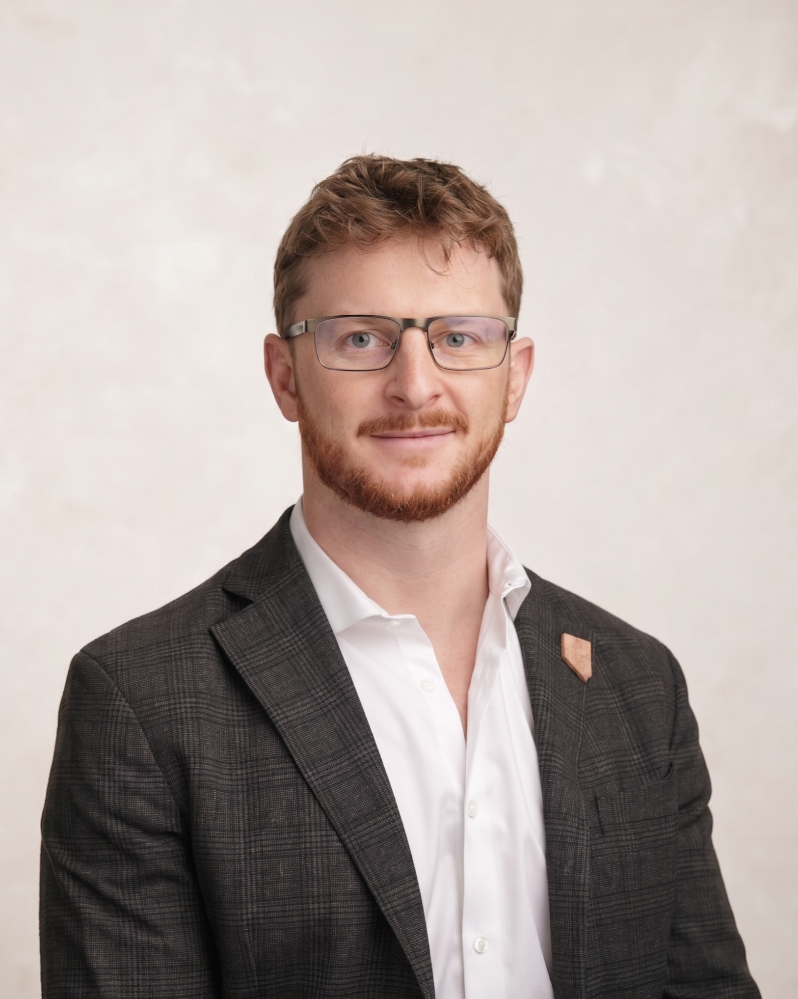 Headshot of Daniel Chafetz, the founder of Apex Consultants, a man with glasses, red hair, red beard, wearing a white shirt and dark suit jacket, and a pin shaped like Nevada made of volcanic tuff, against a light background.