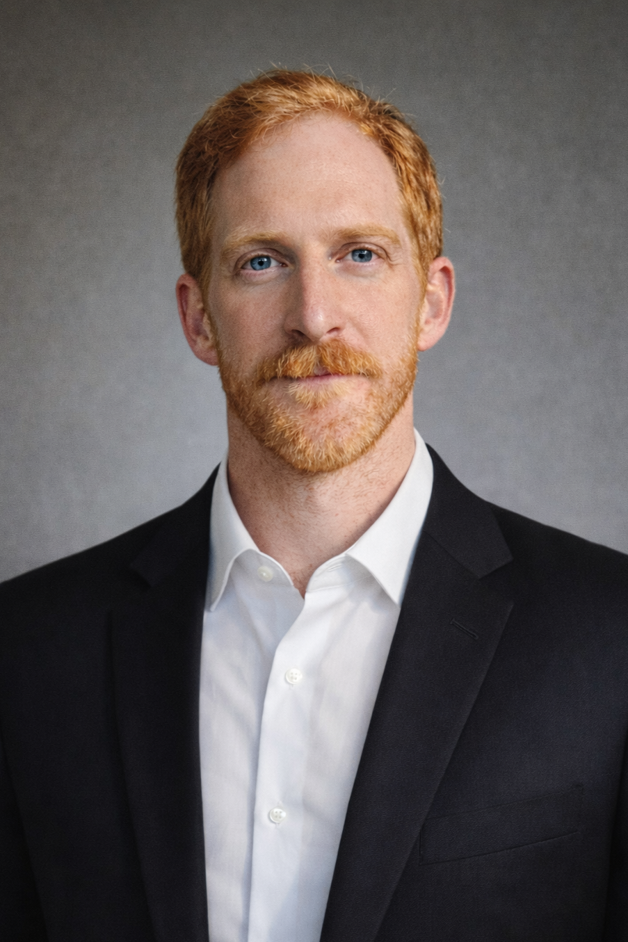 Headshot of Eli DenBesten, a man with red hair and beard, wearing a black suit and white shirt, against a gray background.