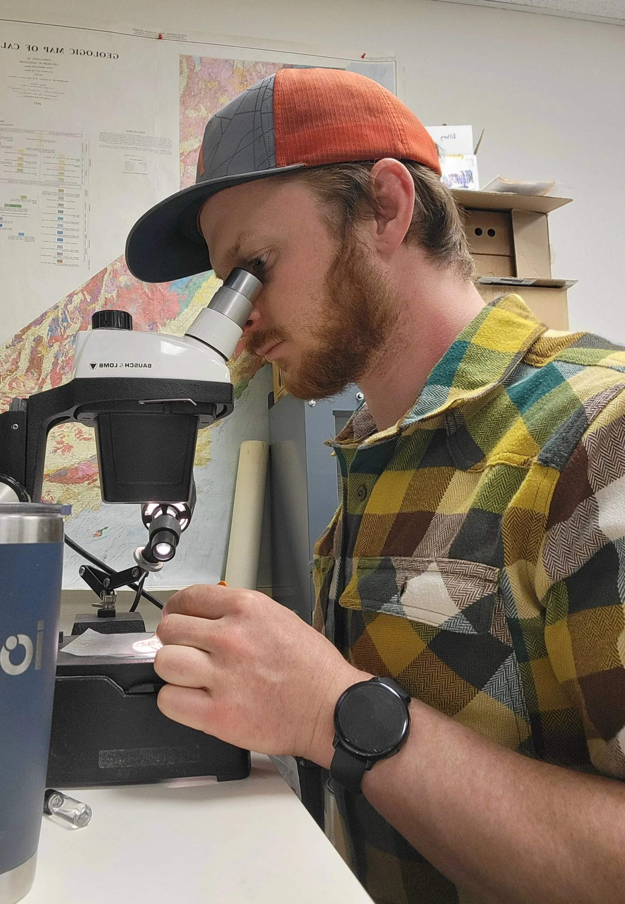 A young man with a beard and colorful checkered shirt is looking into a microscope in a laboratory setting. He is wearing a red and gray cap and a black wristwatch.