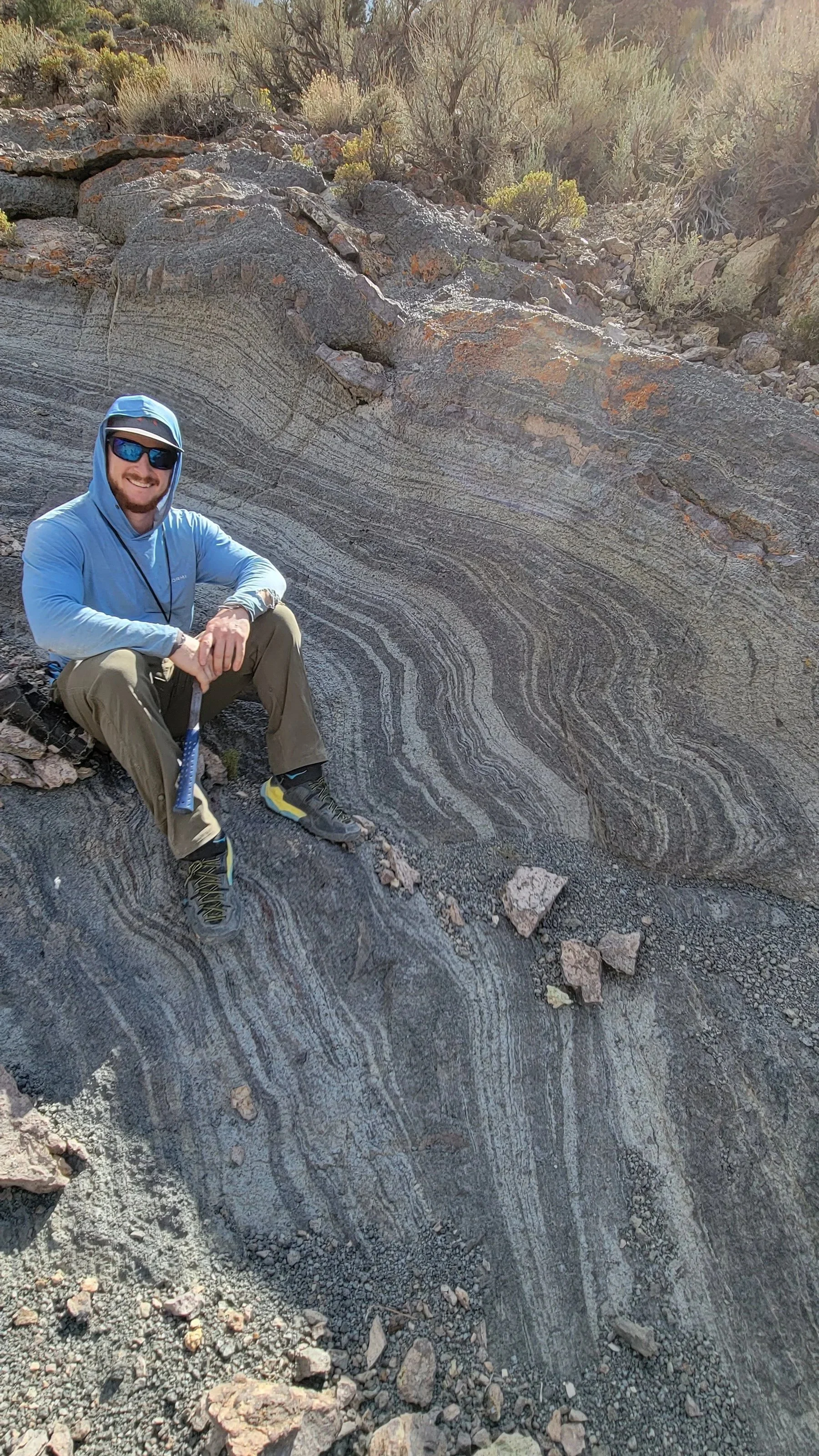 Daniel Chafetz sitting on a beautiful flow-banded rhyolite outcrop in a desert landscape, wearing a blue hoodie, sunglasses, and hiking boots while holding a rock hammer with a hand lens hanging from his neck
