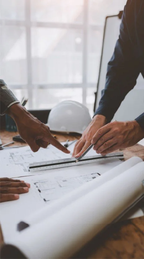Two people review architectural blueprints on a desk, with a hard hat and drawing tools nearby, in a bright office setting.