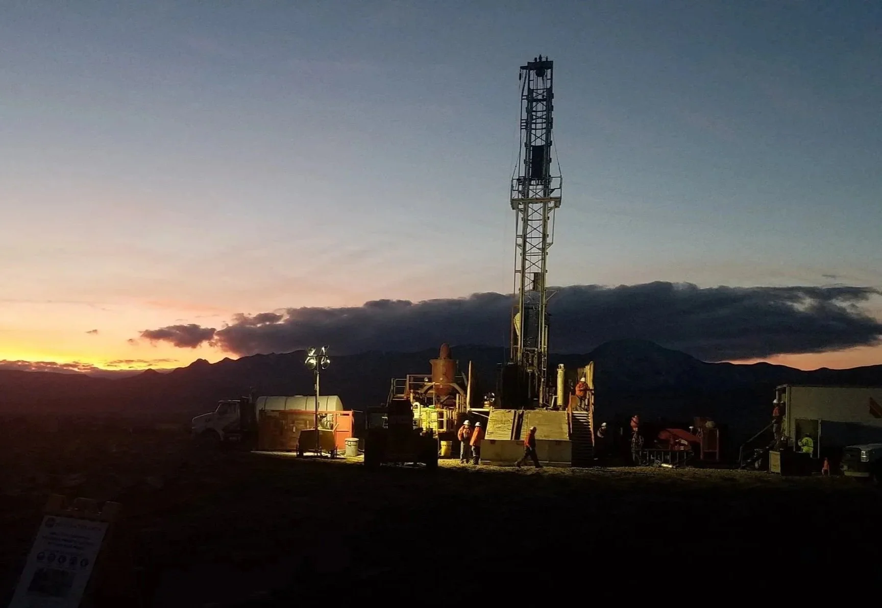 An oil drilling rig at sunset with workers in safety gear, mountains in the background, and equipment illuminated in the foreground.