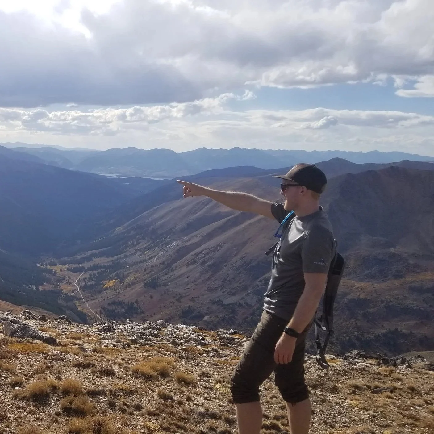 Eli DenBesten with a backpack and hat pointing at distant mountains on a mountain trail under a cloudy sky in Colorado on a summit over 13,000 ft.