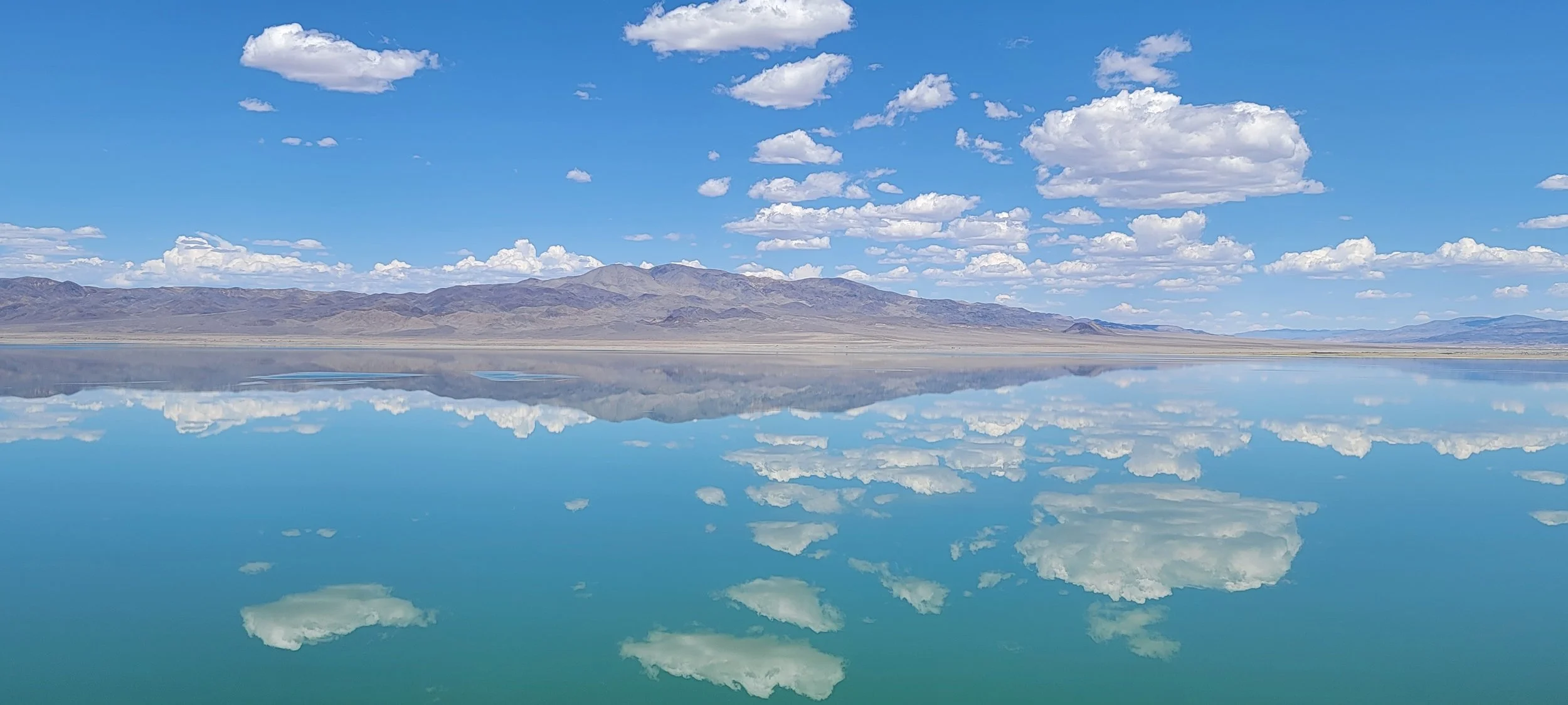 A landscape scene with a clear blue sky, scattered white clouds, and mountains in the distance. The water in the foreground reflects the sky, clouds, and mountains perfectly.