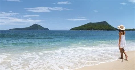 A woman walking on a sandy beach near the ocean with green mountains in the background under a partly cloudy sky in Port Stephens NSW Australia