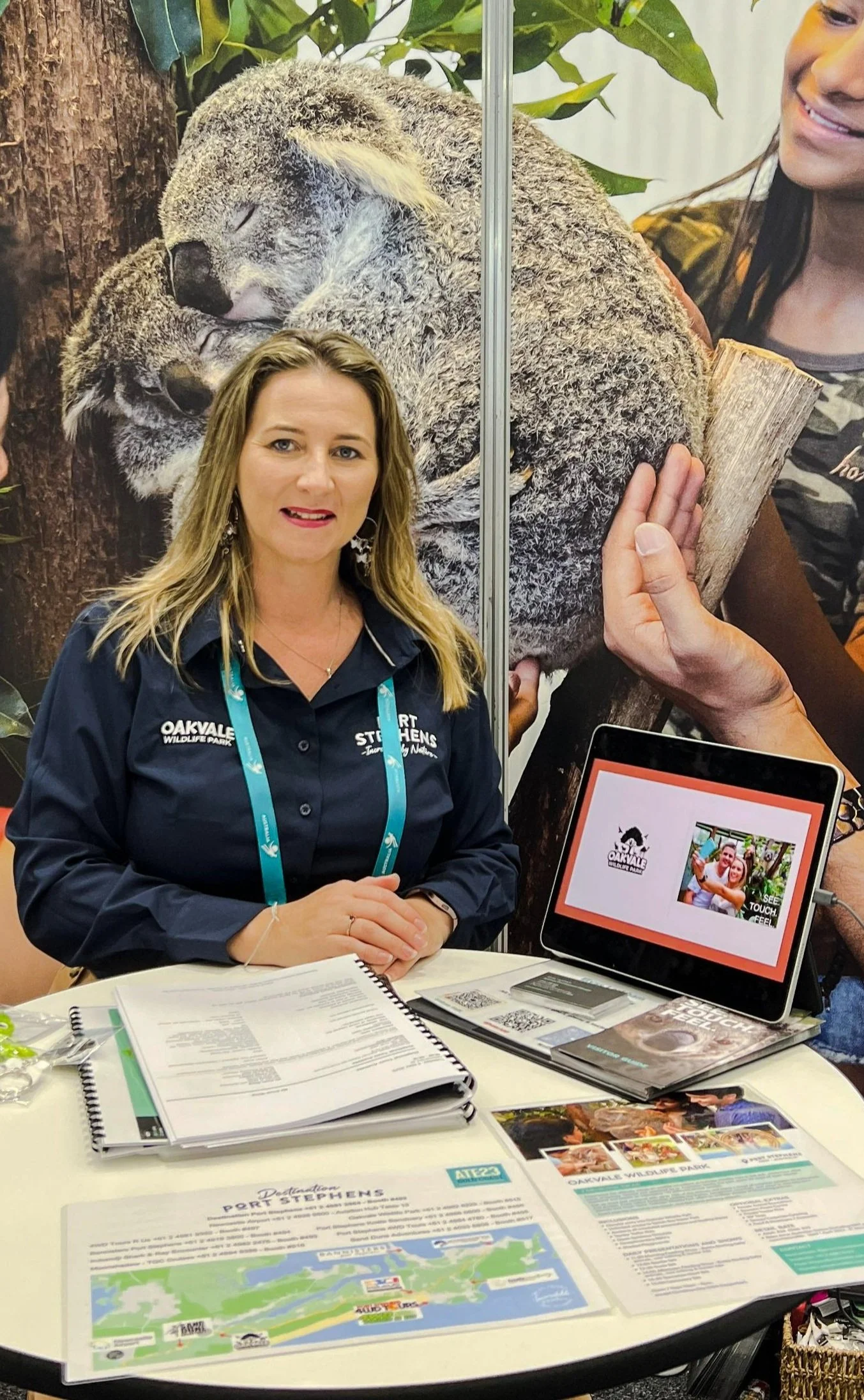 A woman sitting at a desk in front of a large forest-themed poster, with brochures, papers, and a tablet - to show potential clients what is on offer.